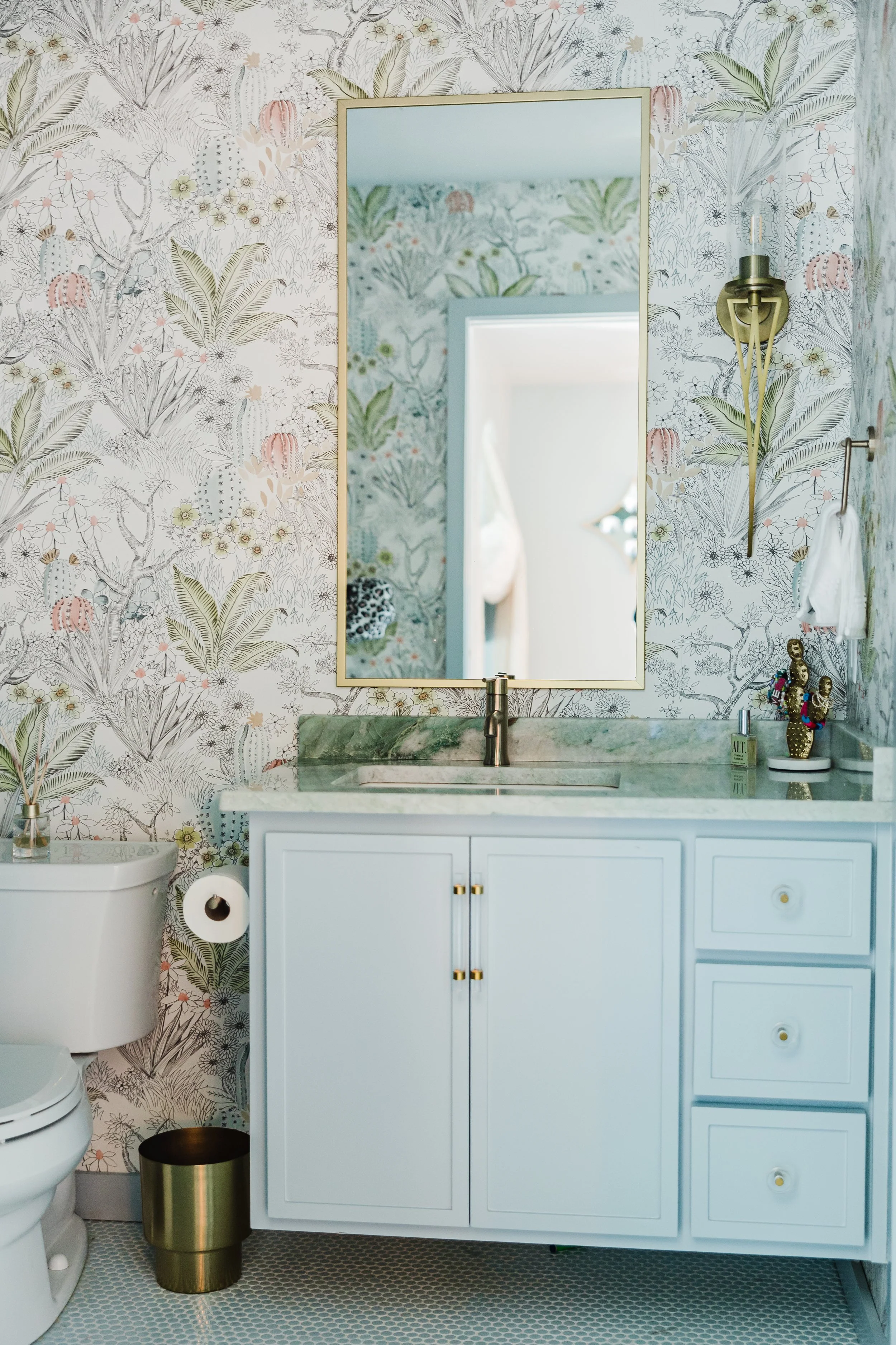 A bathroom with botanical wallpaper, a large mirror, a white vanity with gold accents, and various decorative items including a soap dispenser and figurines.