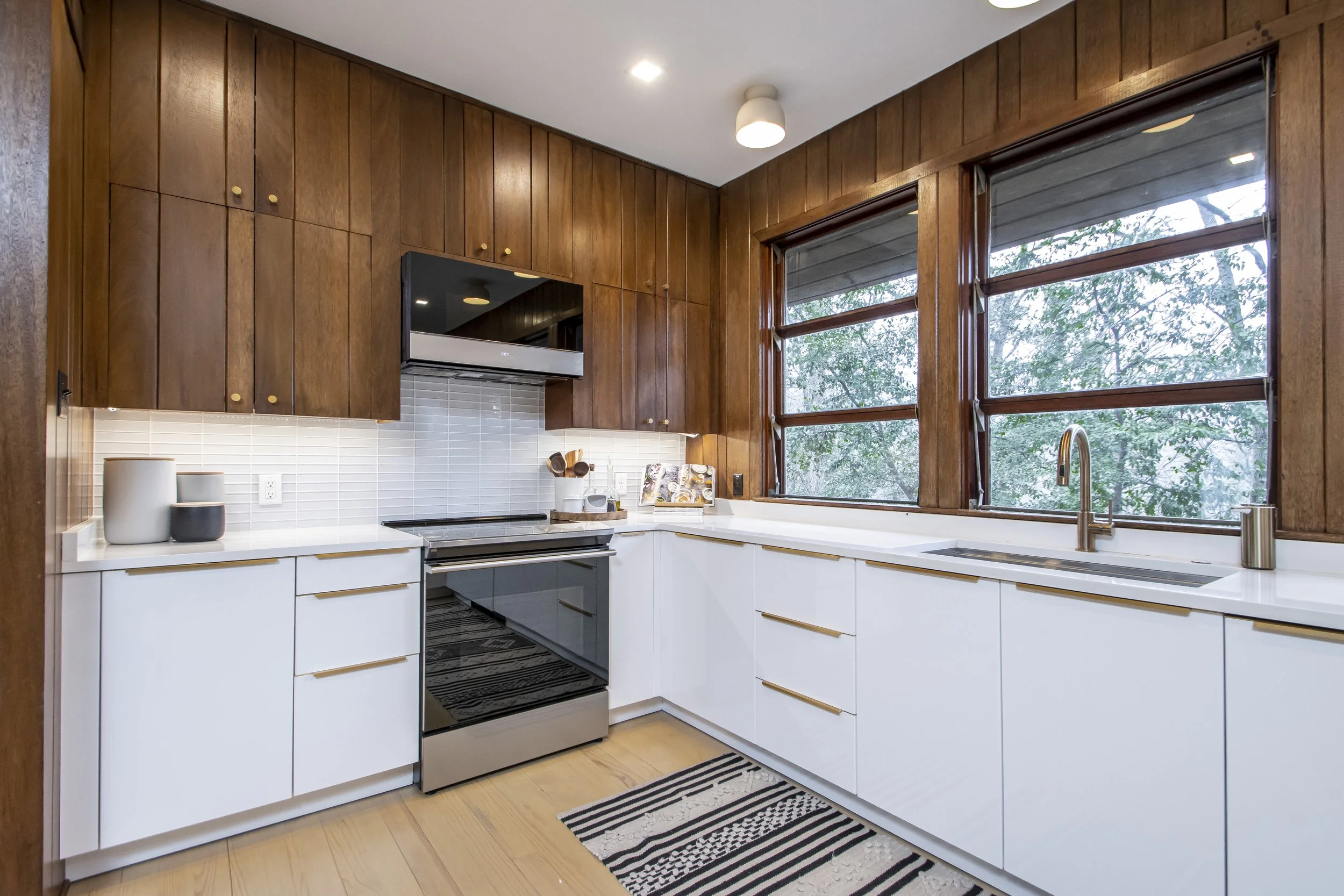 A modern kitchen with white cabinets, a wood-paneled wall, large windows showing green trees outside, a black stove, and small decorative items on the countertop.