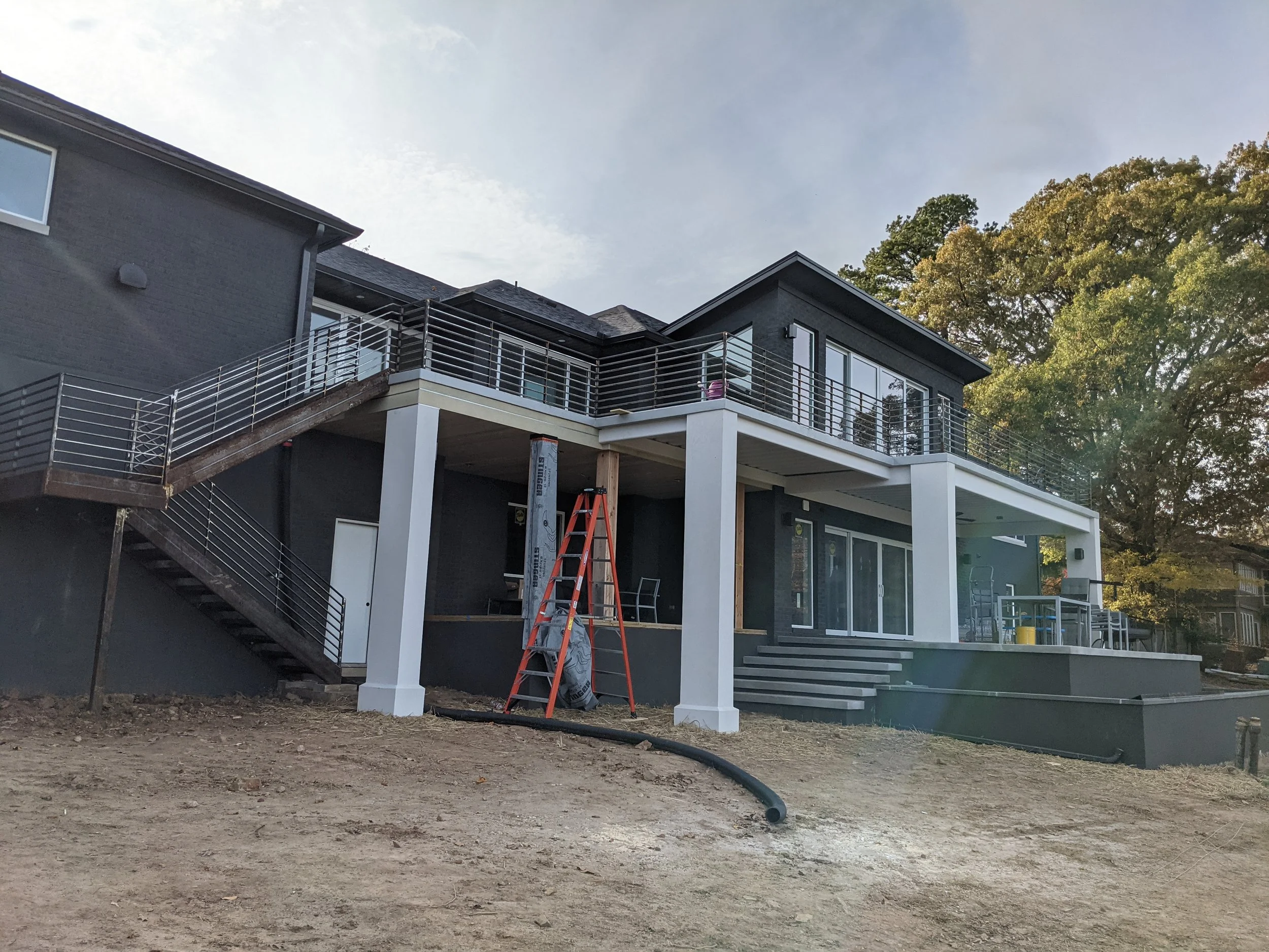 Back deck of a modern house with black exterior walls, white support columns, a metal railing, sliding glass doors, and an outdoor staircase under construction. There is a ladder and construction materials on the ground, and mature trees in the backg