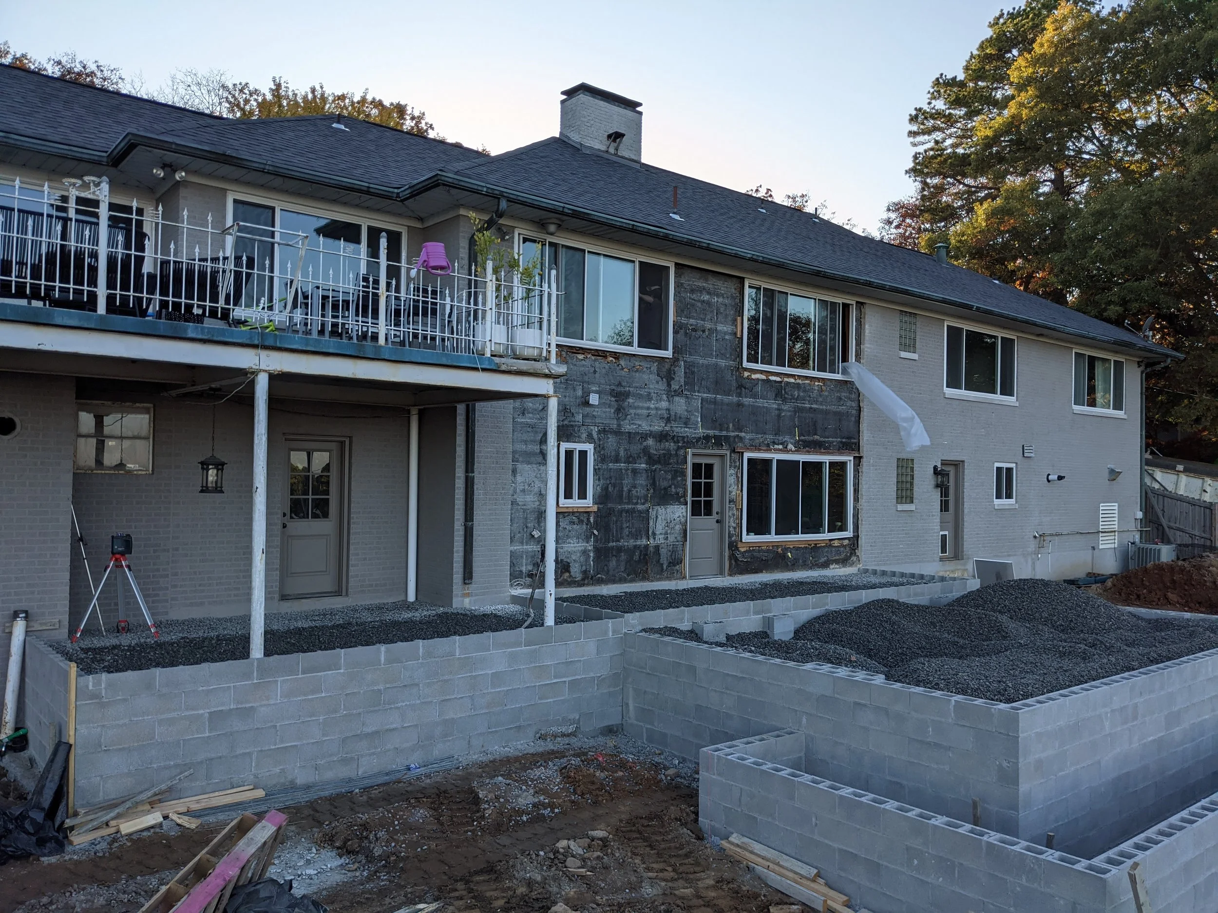 Backyard under construction with cinder block walls, a partially built patio or foundation, and a house with a deck and large windows, surrounded by trees.