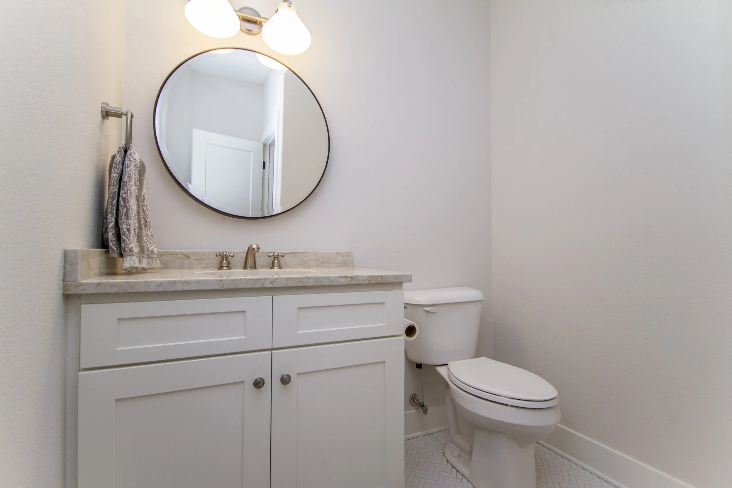 A small bathroom with a white vanity, marble countertop, oval mirror, wall-mounted light fixture, and a white toilet. A beige towel hangs on a silver towel bar to the left of the mirror.