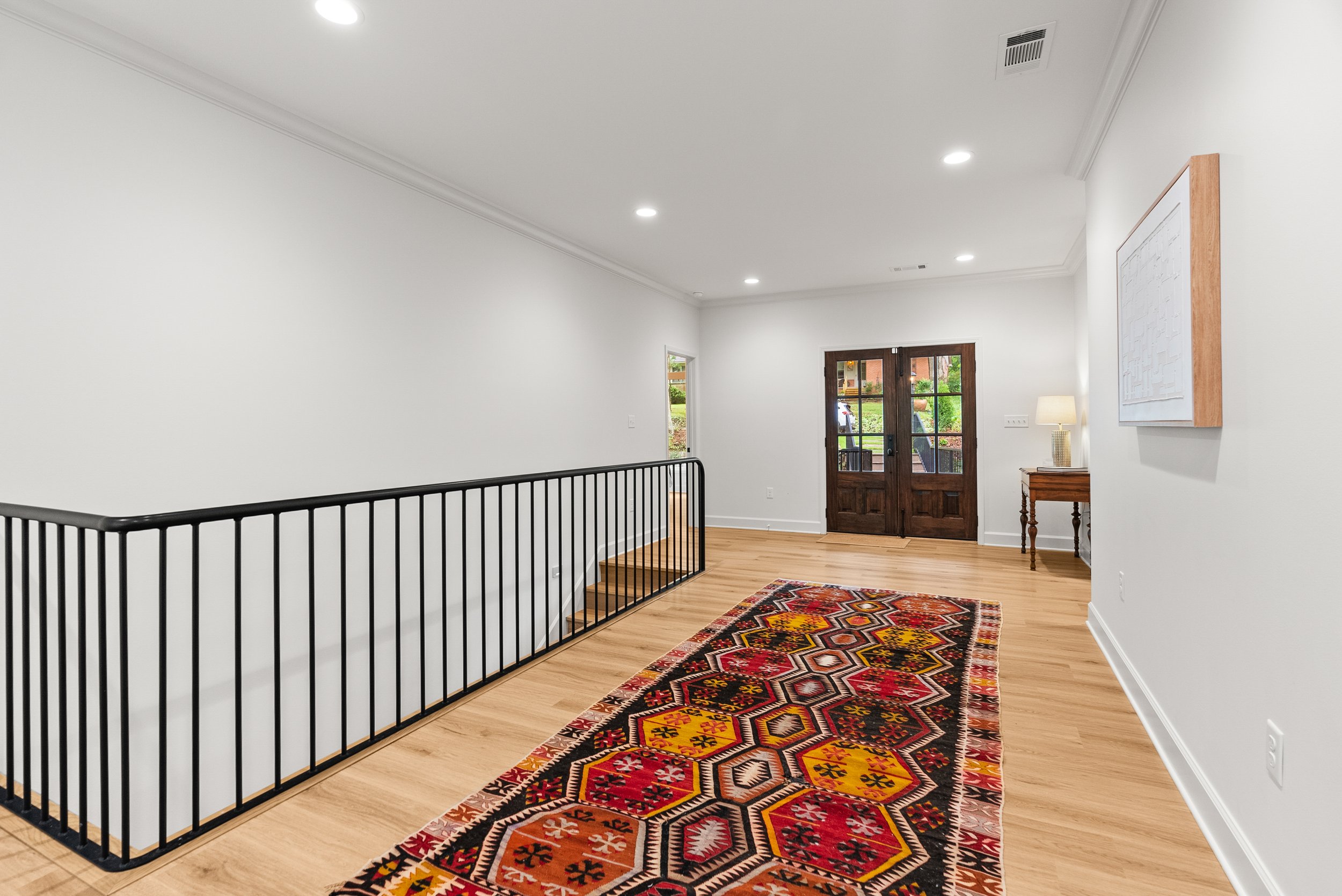 The image shows a spacious hallway with white walls and ceiling, a wooden front door with glass panels, a colorful patterned rug on the light hardwood floor, a small wooden table with a lamp on it, and a black metal railing near a staircase.