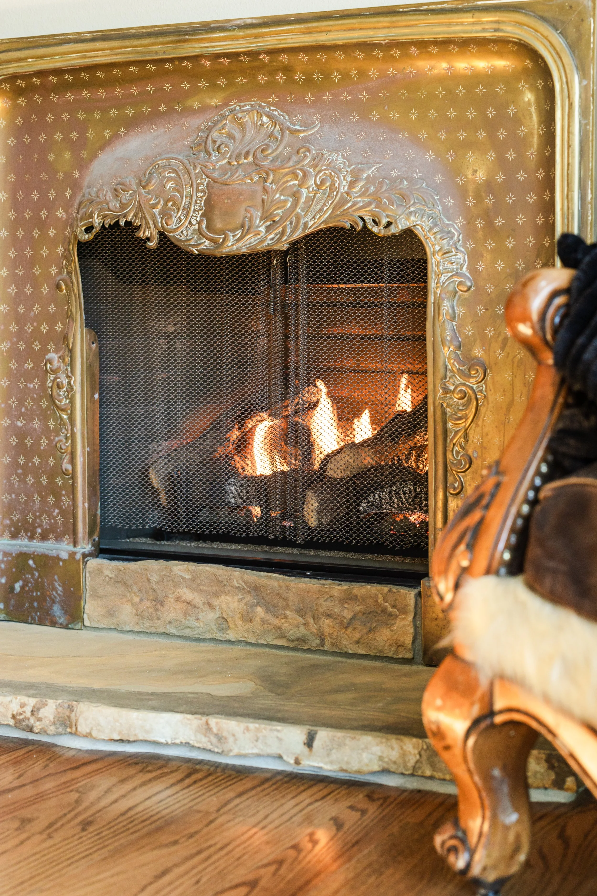 Close-up of a decorated fireplace with a fire burning inside. The fireplace has ornate gold-colored trim and a mesh screen. A wooden chair with curved armrest is partially visible on the right side of the image. The fireplace is set on a stone hearth