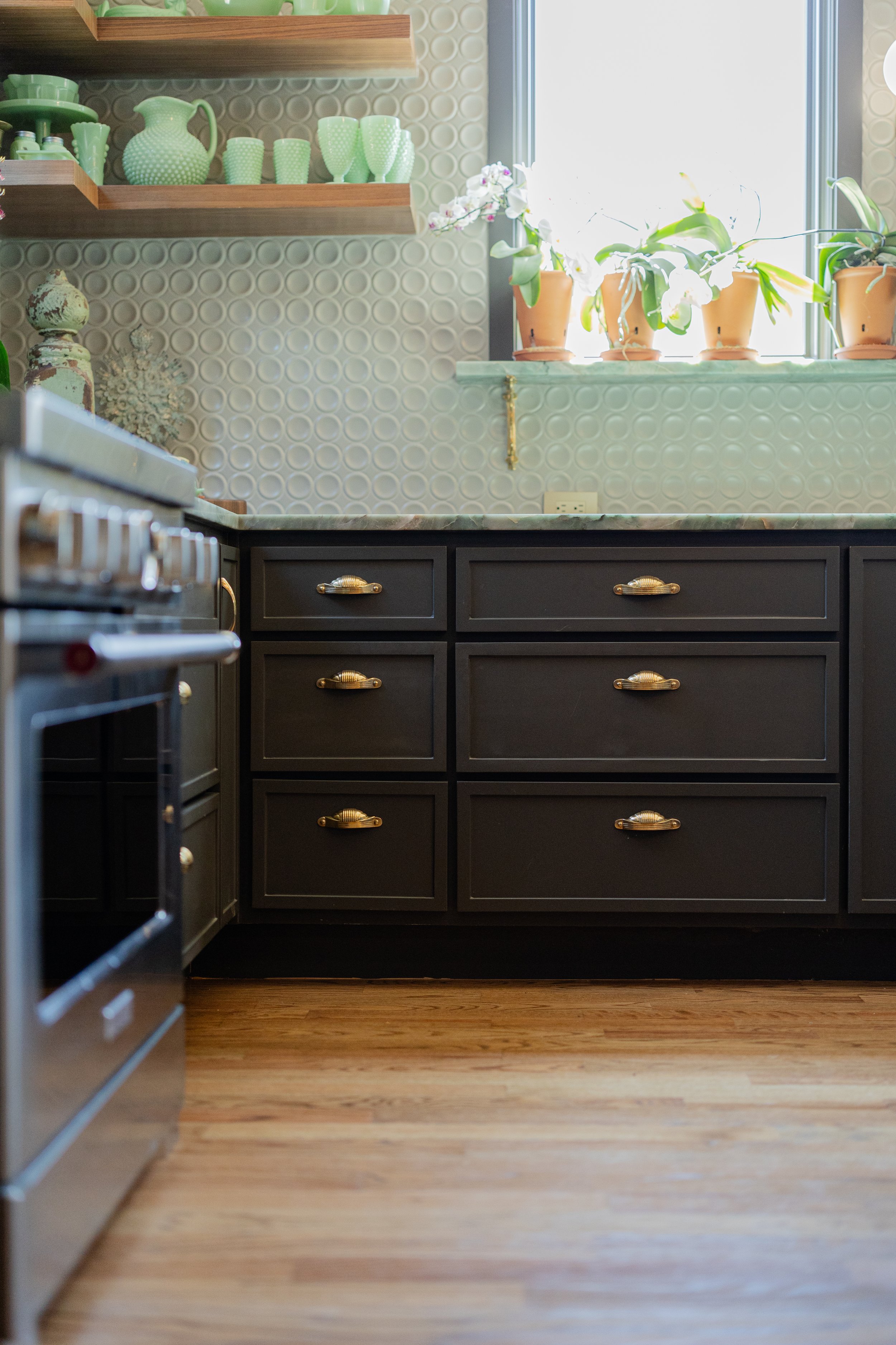Kitchen interior with black cabinets, wooden flooring, and shelves with green dishware and vases; white orchids on the windowsill, sunlight streaming through the window.