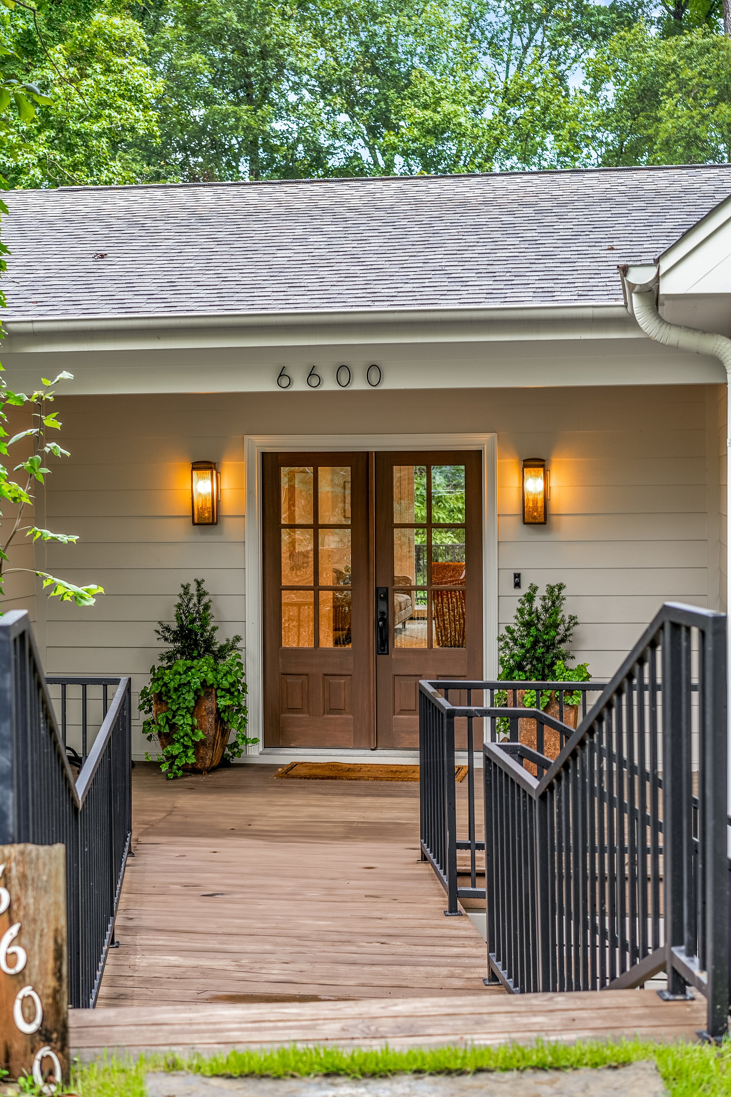 Front entrance of a house with a wooden porch, brown door with glass windows, two black lantern-style wall lights, potted plants on either side of the door, and house number 6600 displayed above the door, surrounded by trees and greenery.