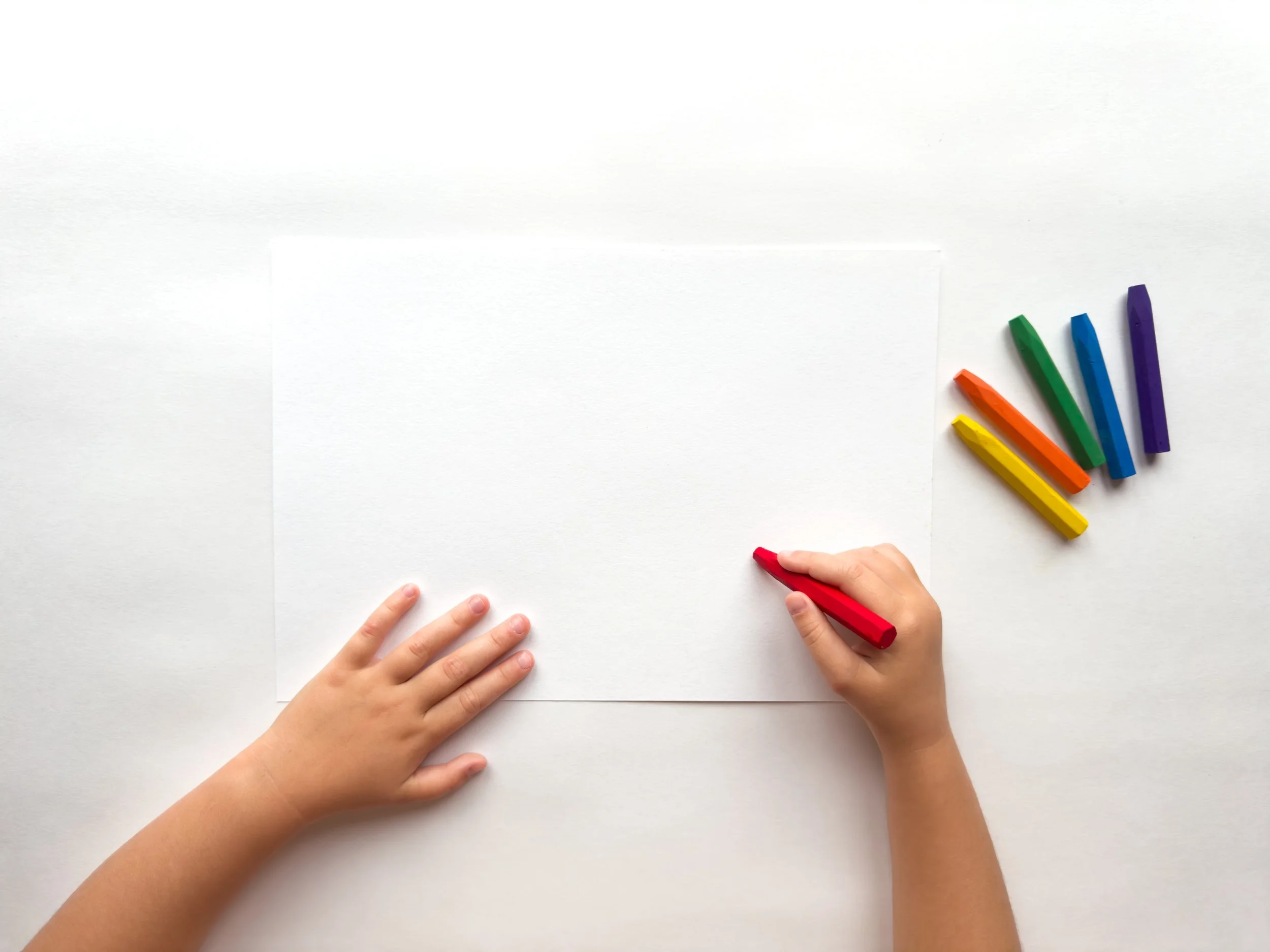 A child's hands holding a red crayon over a white sheet of paper on a white surface, with a rainbow-colored crayon set to the right.