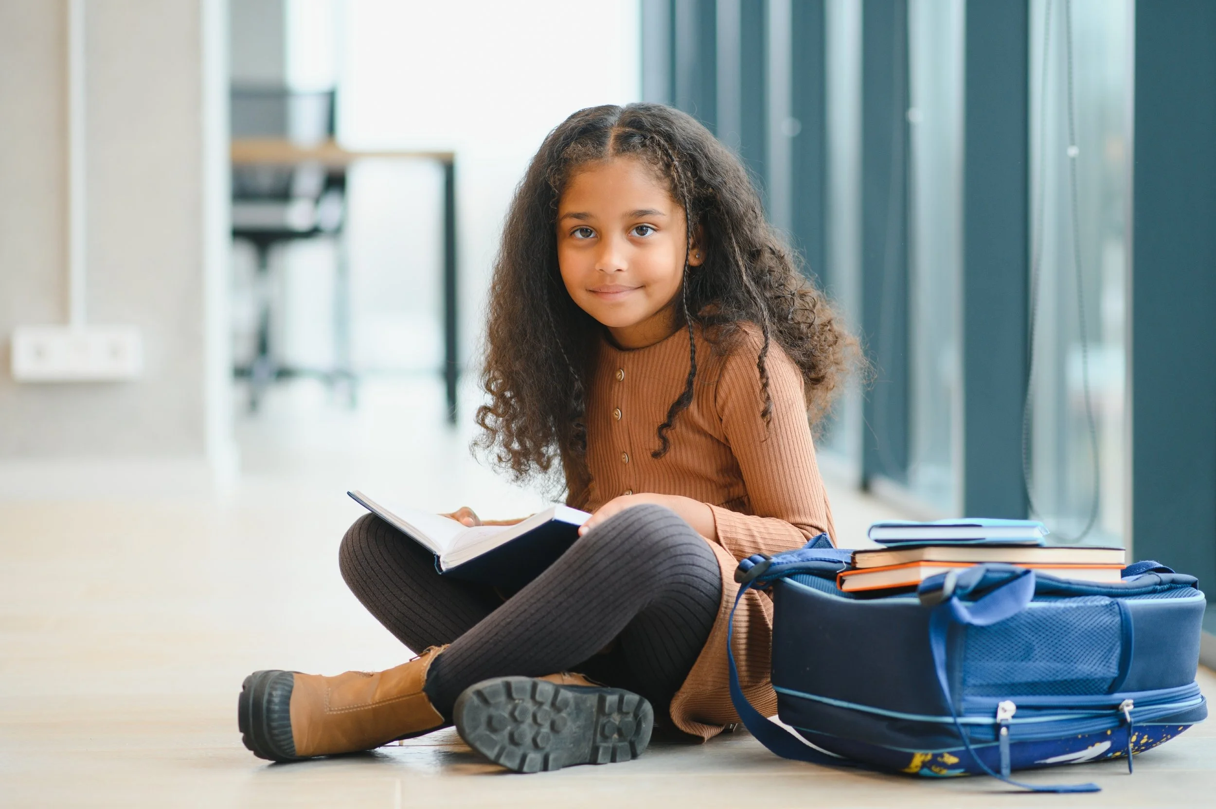 A young girl with curly hair, sitting on the floor, smiling, holding an open book with stacked books and a blue backpack nearby, in a bright modern indoor setting.