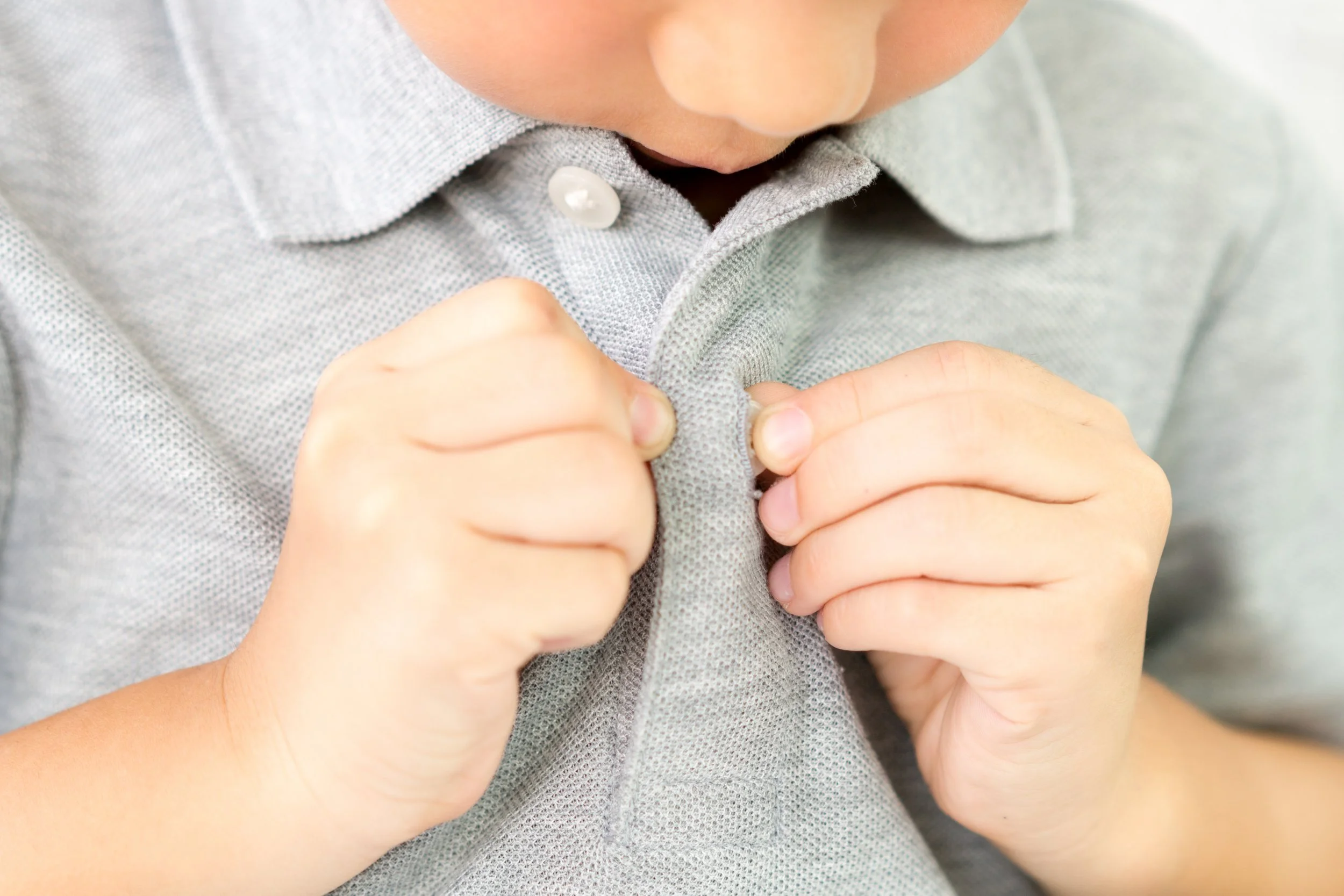 Close-up of a young child buttoning a grey shirt, with focus on the child's hands and chin.