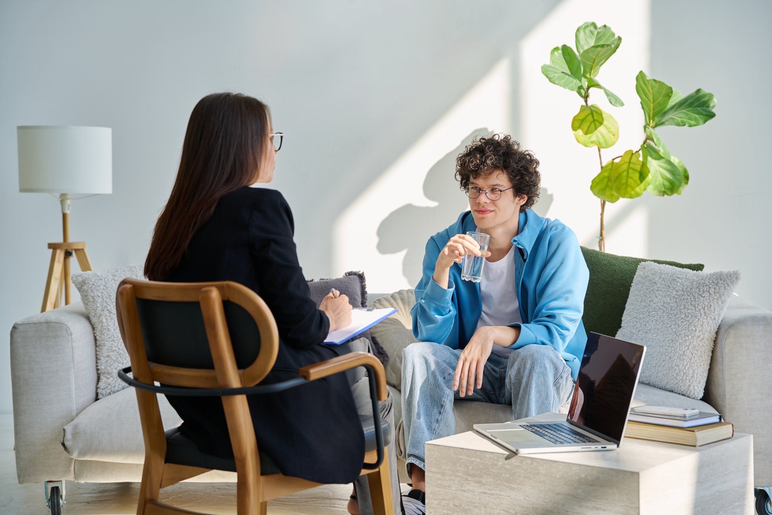 A young man with curly hair and glasses sitting on a couch, drinking water, speaking with a woman holding a notepad, in a bright modern living room.