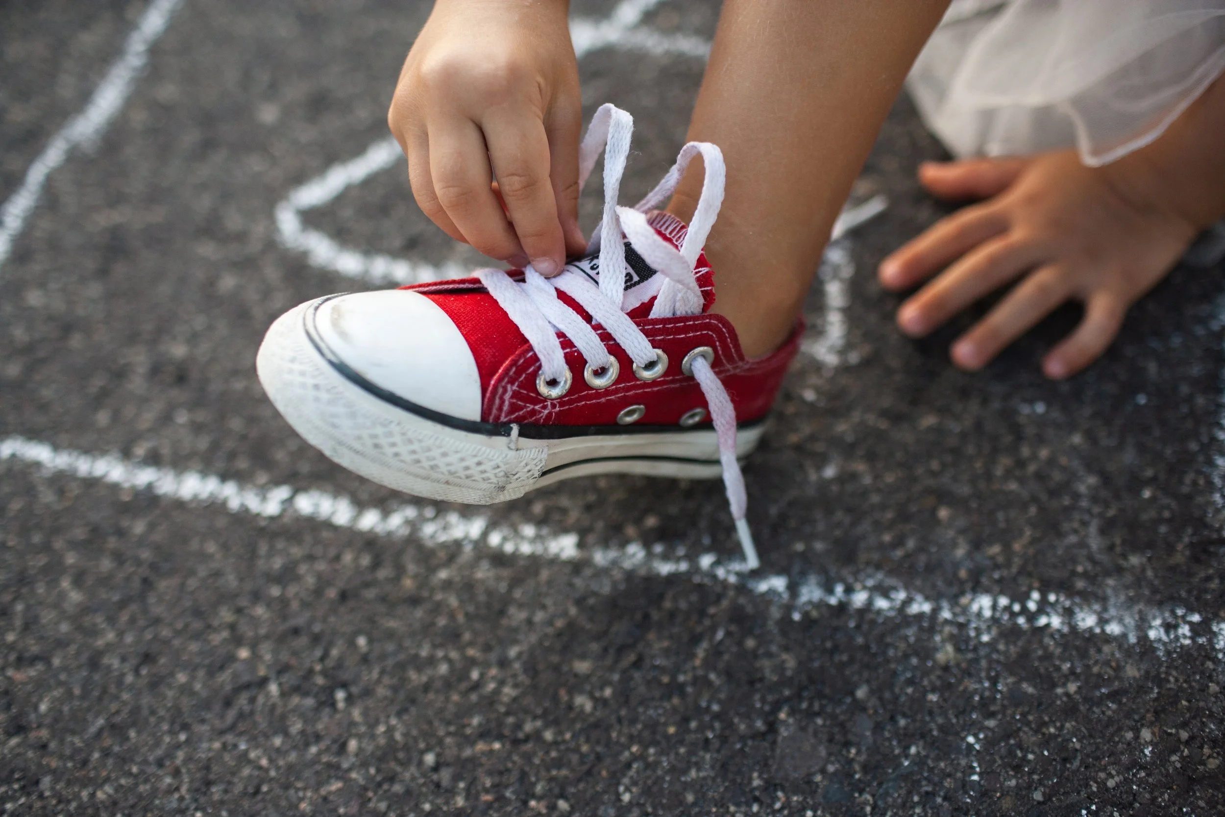 Child tying shoelaces on a red sneaker on asphalt, with white chalk lines on the ground.