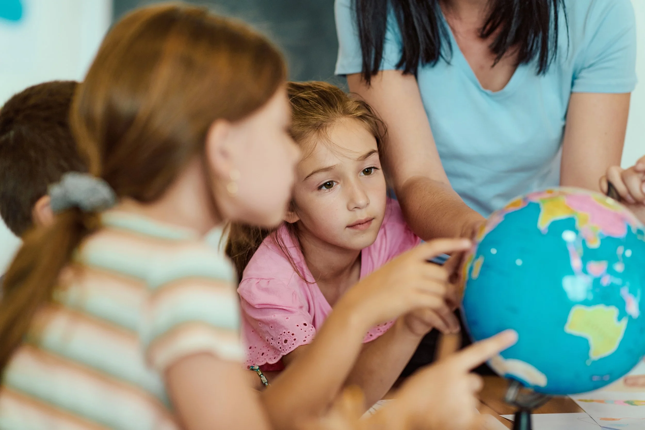 A group of children, including a girl in a pink shirt, and an adult woman wearing a light blue shirt, gathered around a colorful globe, pointing and learning about geography.