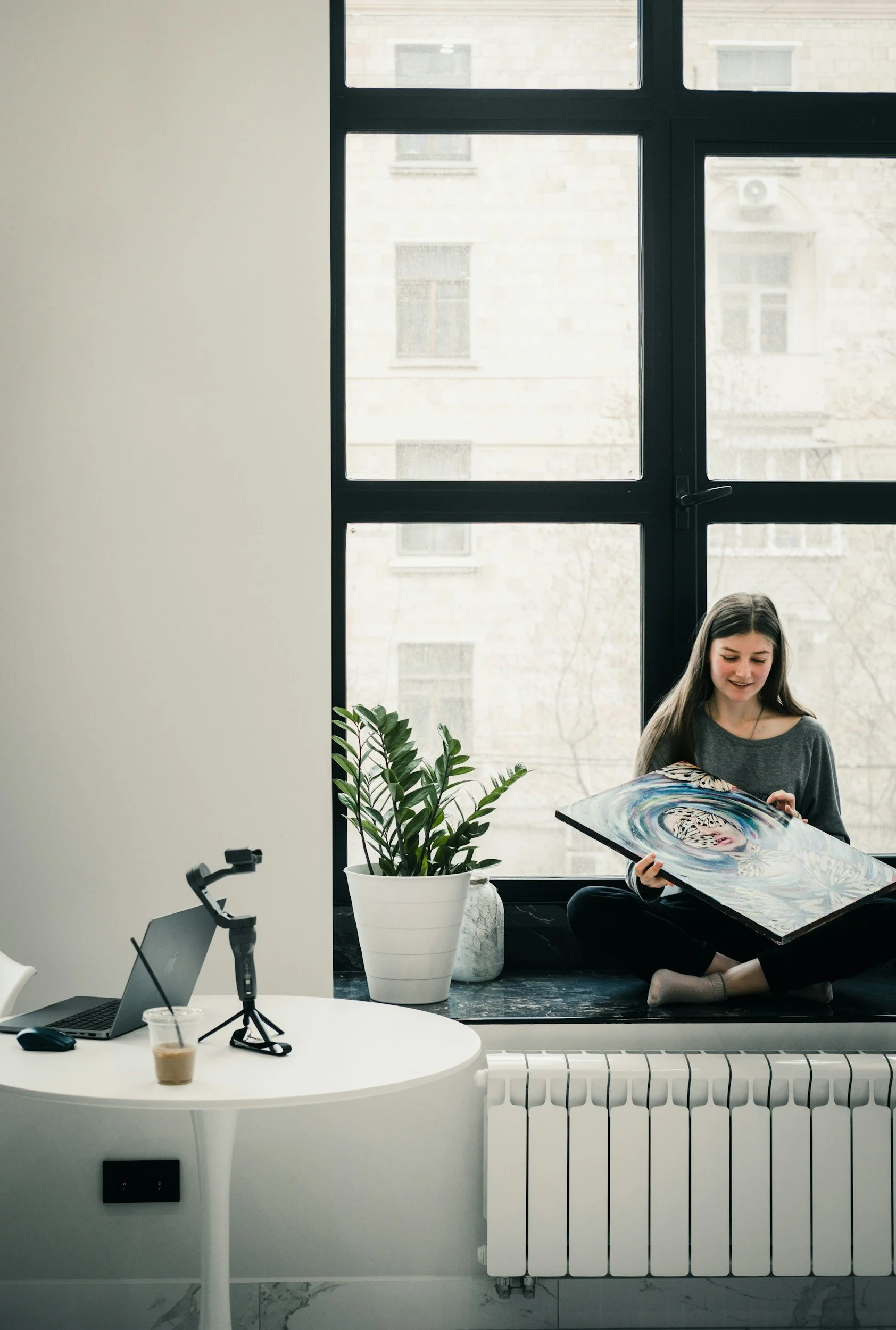 A young woman with long dark hair sitting on a black marble window sill, holding and looking at a colorful swirl abstract art piece. There is a large window behind her, a potted plant on her left, and a building with stone walls visible outside.