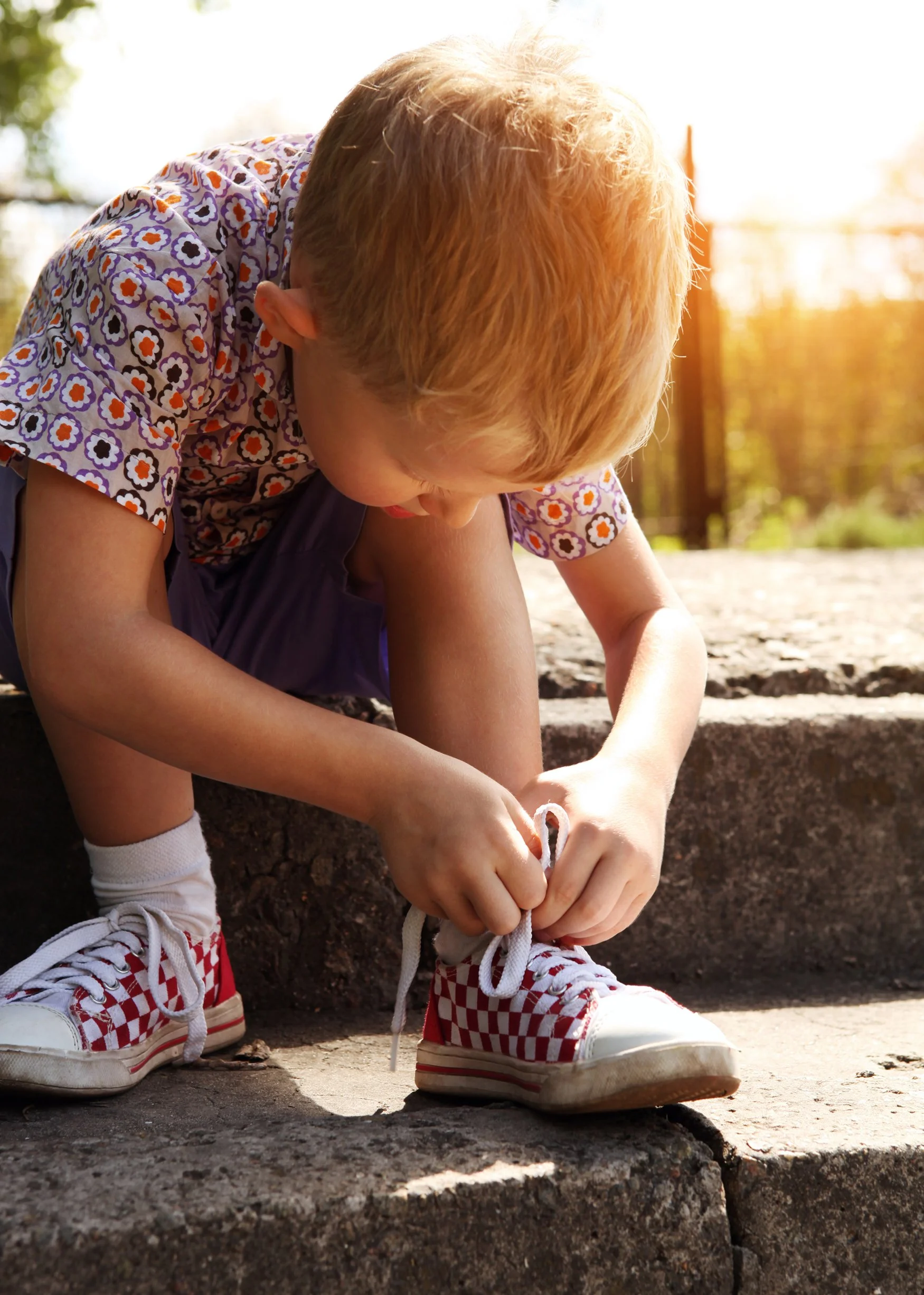 A young child with red hair, wearing a patterned shirt with orange, purple, and white flowers, red and white checkered shoes, and white socks, tying the laces of their shoes while sitting outdoors in the sunlight.