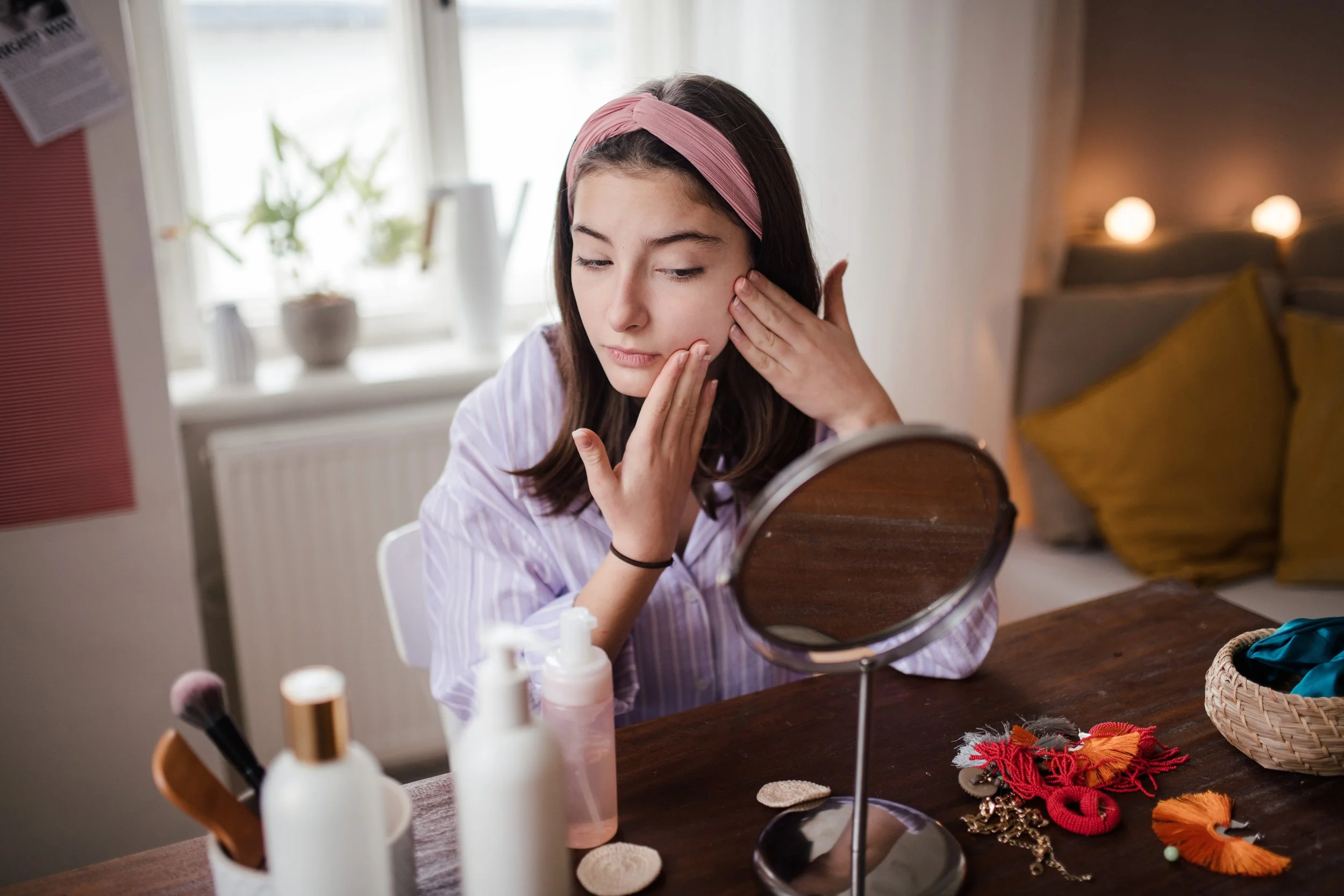 A young woman with long dark hair wearing a pink headband looks into a mirror while touching her face, sitting at a table with skincare bottles, makeup brushes, and handmade accessories nearby.