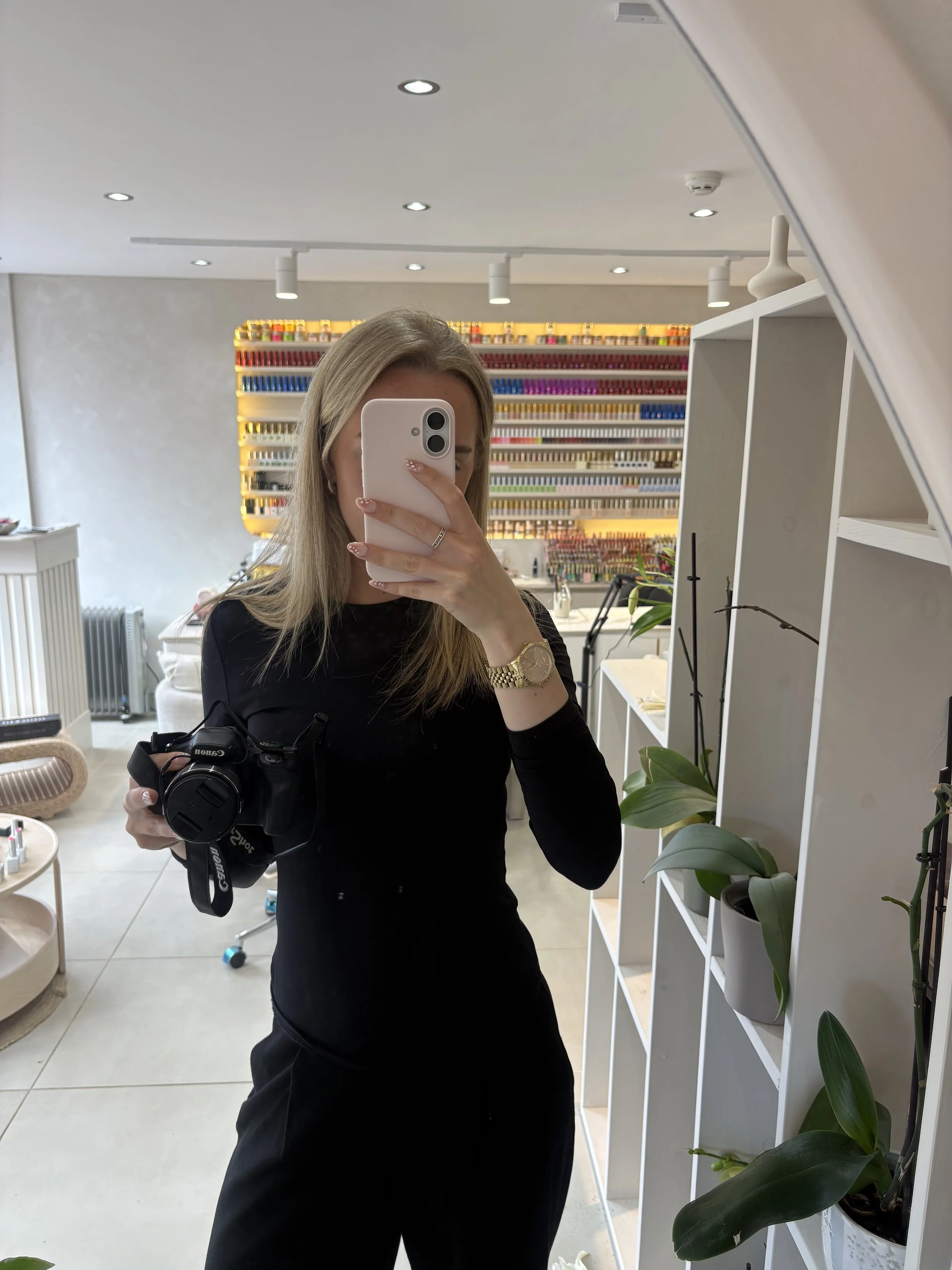 A woman taking a mirror selfie in a store or salon, holding a camera and wearing a black top, with colorful nail polish samples displayed on a wall behind her.