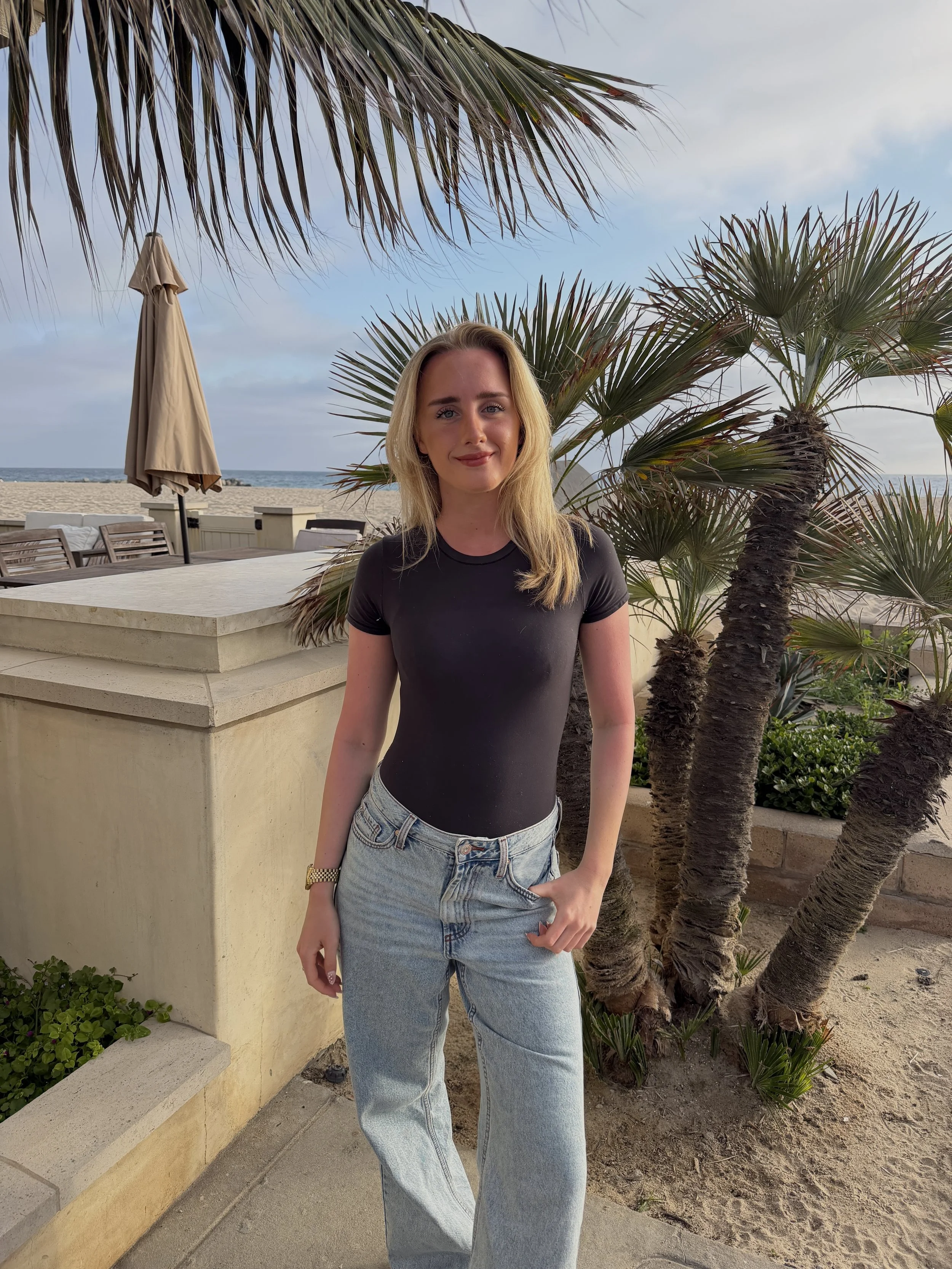 A woman with blonde hair wearing a black t-shirt and light blue jeans standing outdoors near palm trees, with the beach and ocean visible in the background.