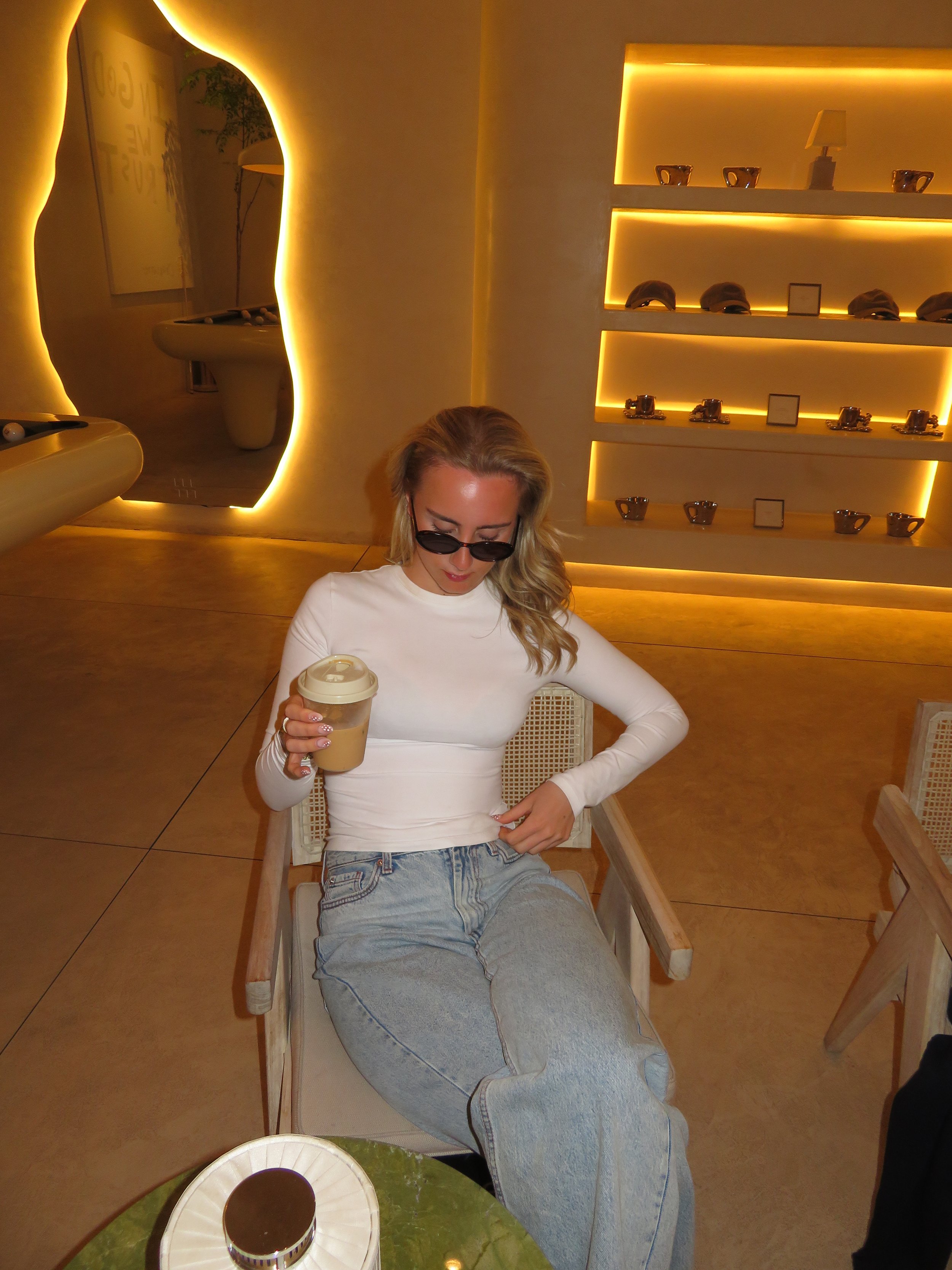 A woman with blonde hair wearing sunglasses, a white long sleeve shirt, and jeans, sitting in a chair holding a coffee cup, in a room with modern lighting and shelves displaying ceramics and decorative items.