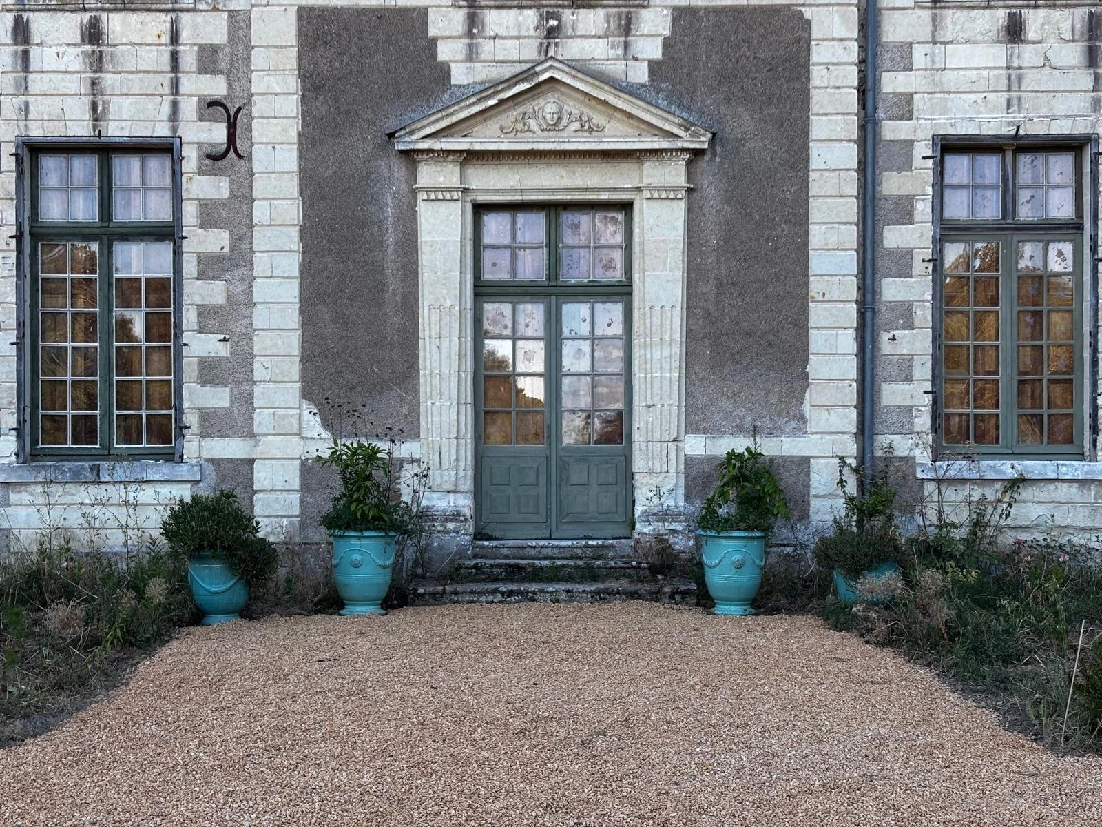 Façade d'une maison ancienne avec deux fenêtres et une porte. La maison est en pierre avec des volets verts, et il y a des plantes en pot turquoise devant la porte.
