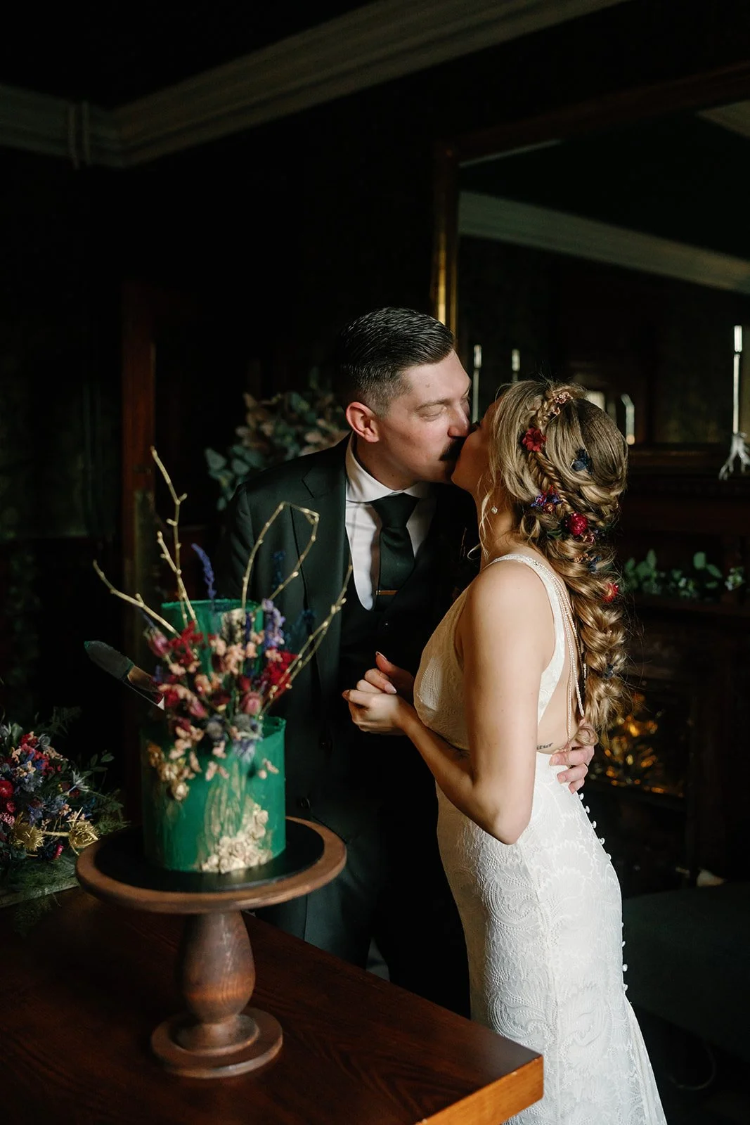 Newly married couple sharing a kiss beside their contemporary wedding cake decorated with colourful dried flowers, emerald and gold detailing.