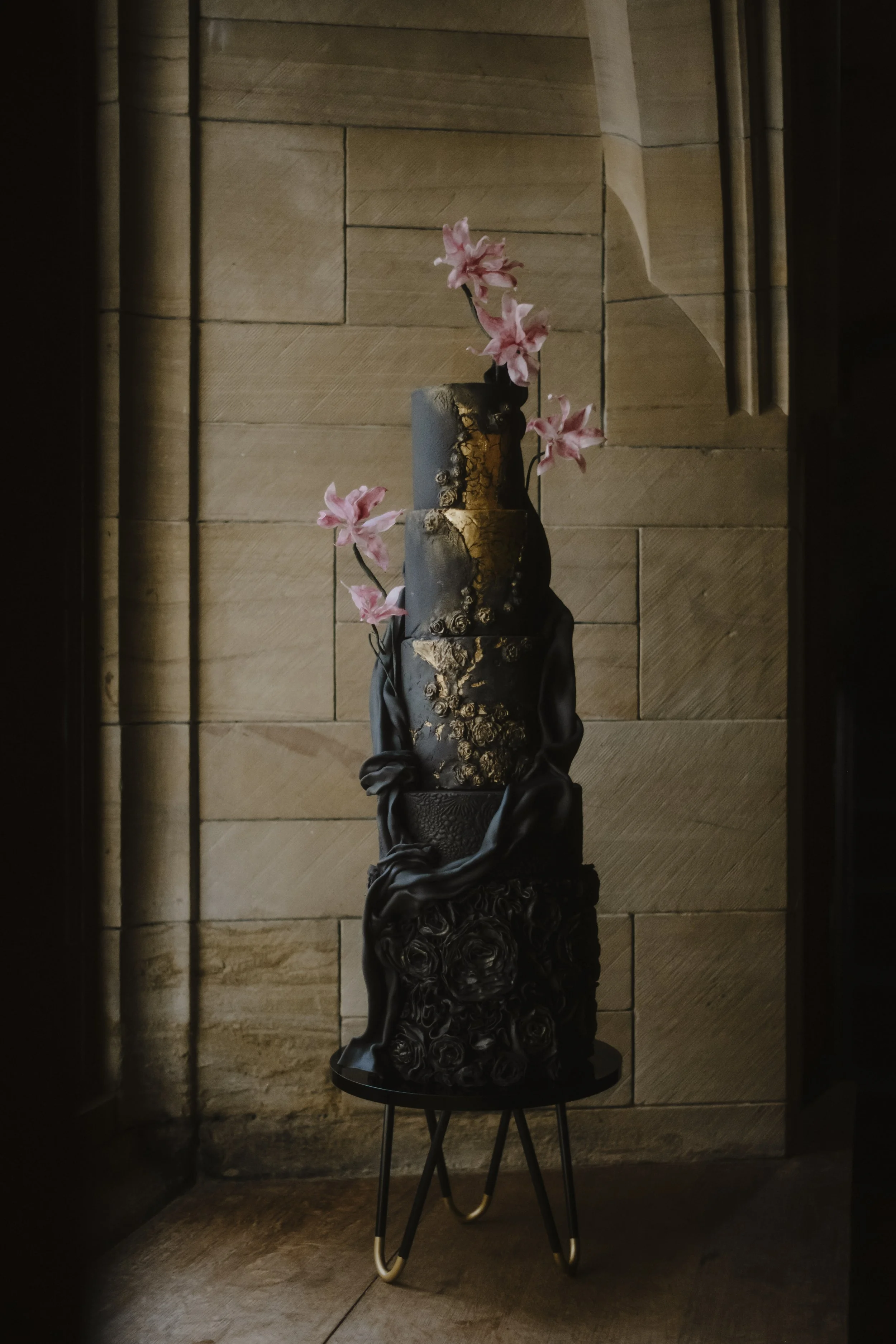 Dramatic black multi-tier luxury wedding cake with gold detailing, flowing fondant and handmade burgundy wafer paper flowers displayed against a stone backdrop at Achnagairn Castle
