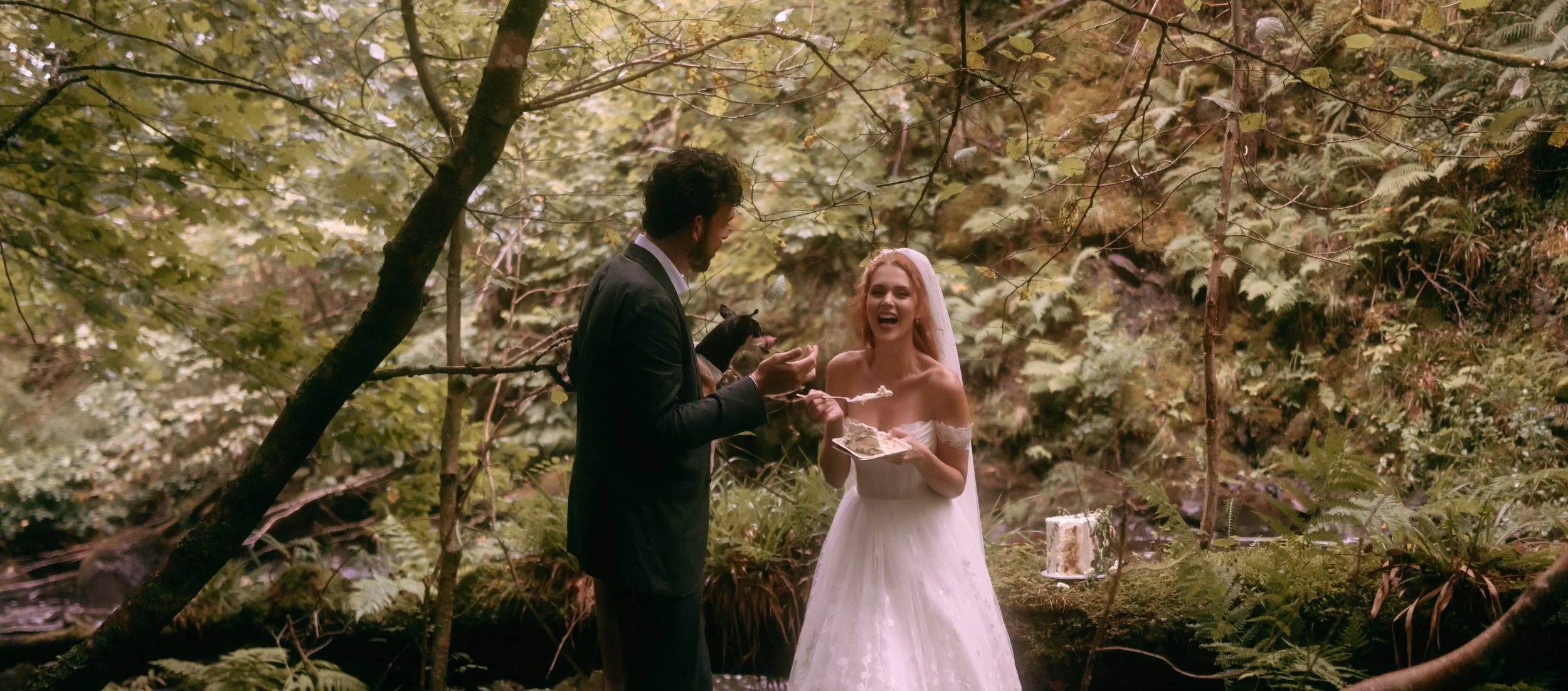 Bride and groom laughing together as they enjoy their wedding cake during an intimate woodland wedding celebration.
