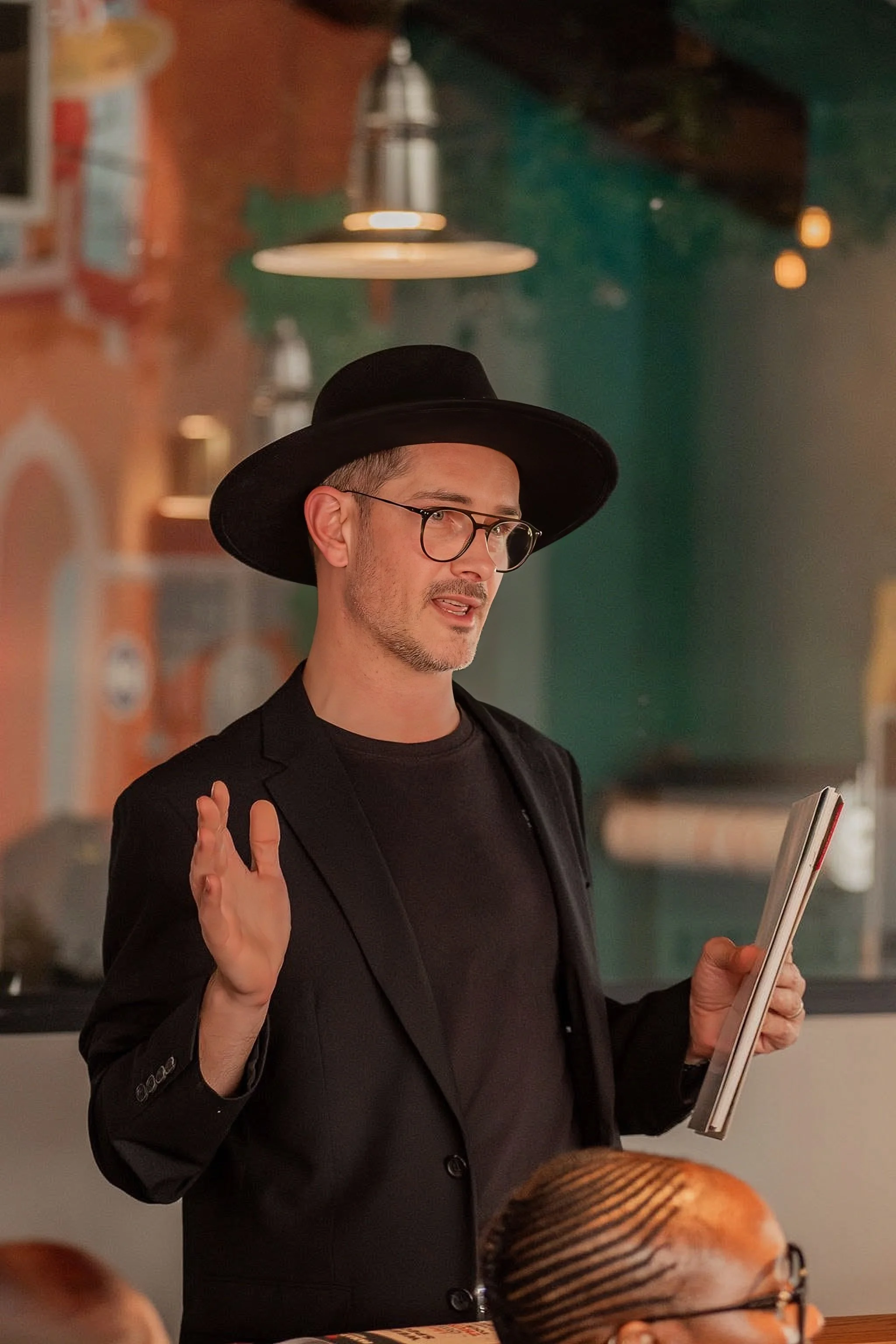 A man wearing glasses, a black hat, and a black suit is gesturing with his right hand while holding a drawing pad in his left hand. He appears to be speaking or explaining something in a setting that resembles a cafe or casual restaurant.