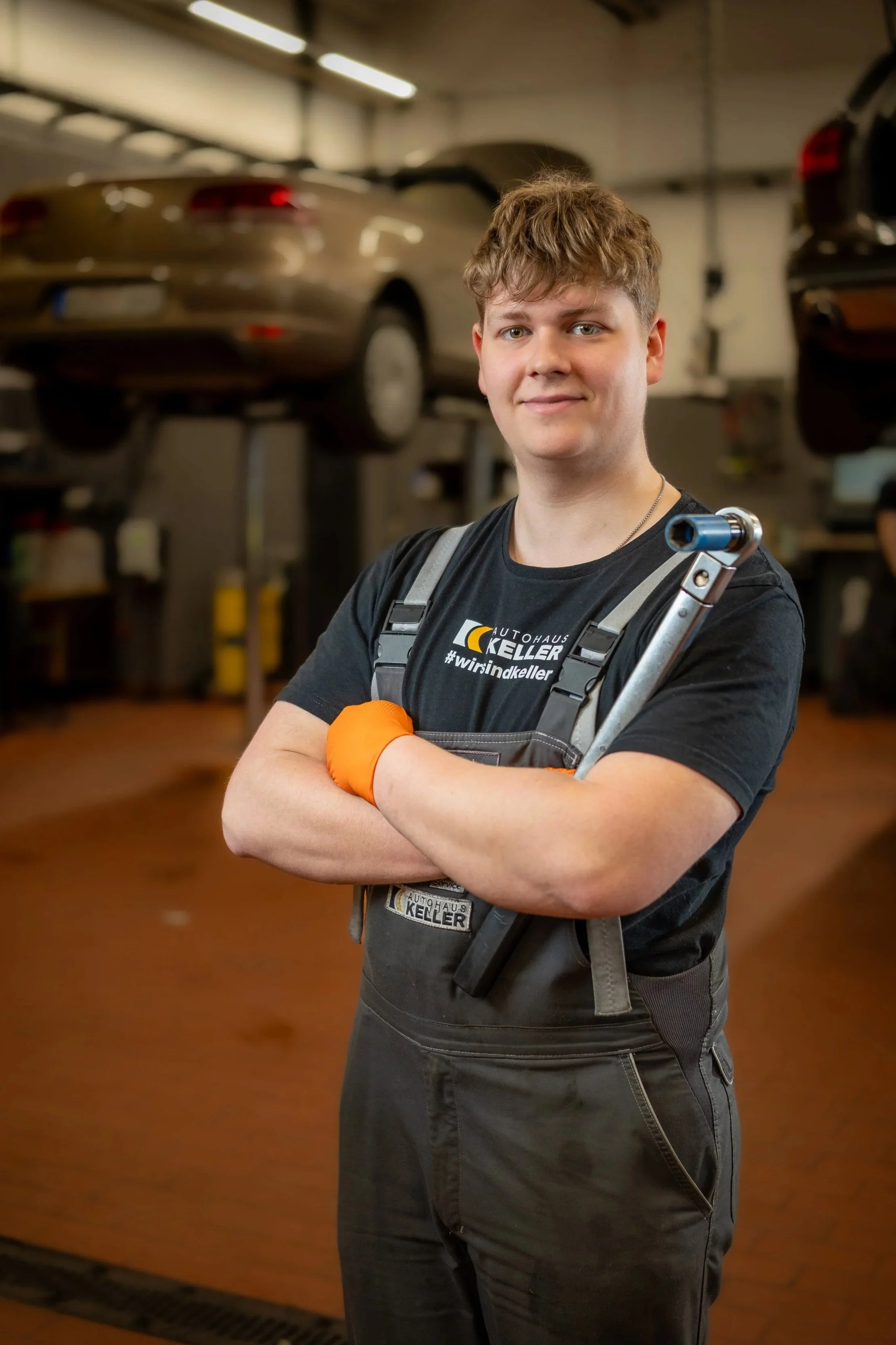 Ein junger Mann steht in einer Werkstatt, trägt schwarze Arbeitskleidung mit einem Firmenlogo, trägt eine orange Arbeitshandschuh am rechten Arm und hält einen Schraubenschlüssel. Er lächelt die Kamera an.