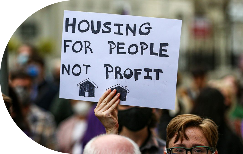 A photograph depicting a person holding a sign saying “Housing For People, Not Profit”.