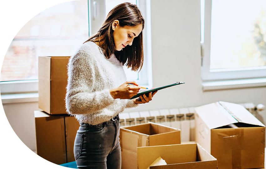 A photograph depicting a woman looking at a clipboard while standing in a room full of cardboard packing boxes.