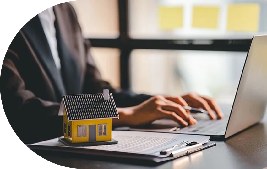 A photograph depicting a person typing on a laptop while a miniature-sized house lies on a stack of papers.
