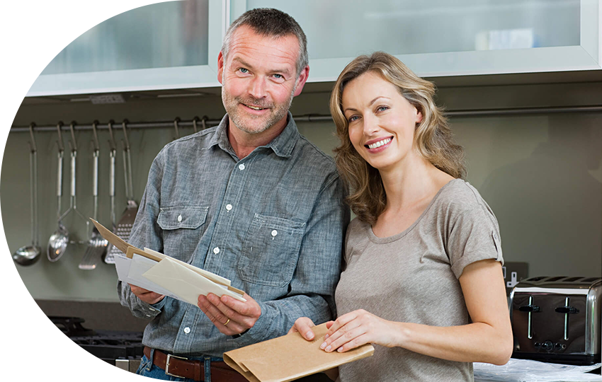 A photograph depicting a middle-aged couple standing in a kitchen while holding a stack of papers and documents.
