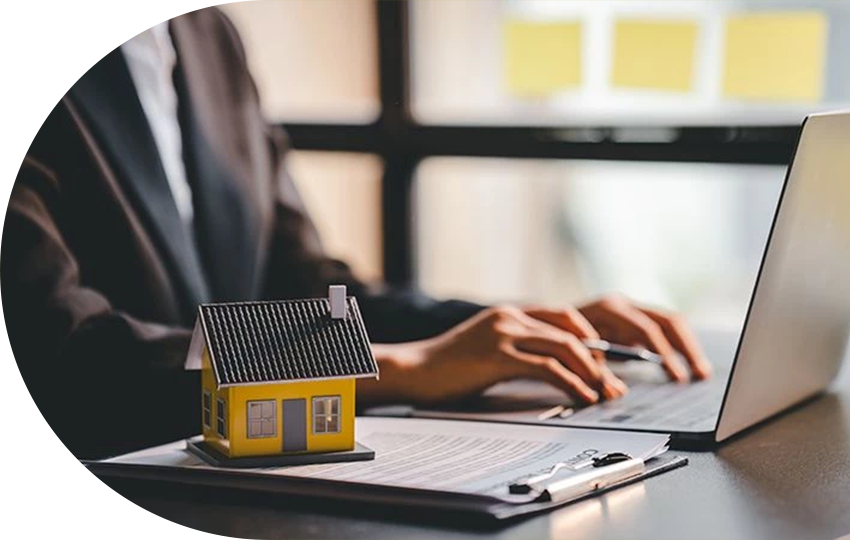 A photograph depicting a person typing on a laptop while a miniature-sized house lies on a stack of papers.