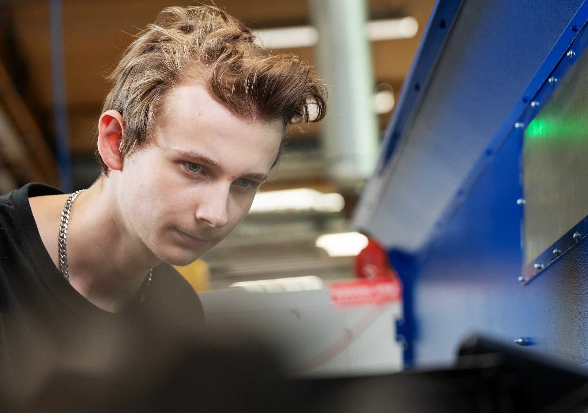 En ung man med ljus hud, kort lockigt hår tittar fokuserat på en cnc-svarv utanför bildens vänstra sida.