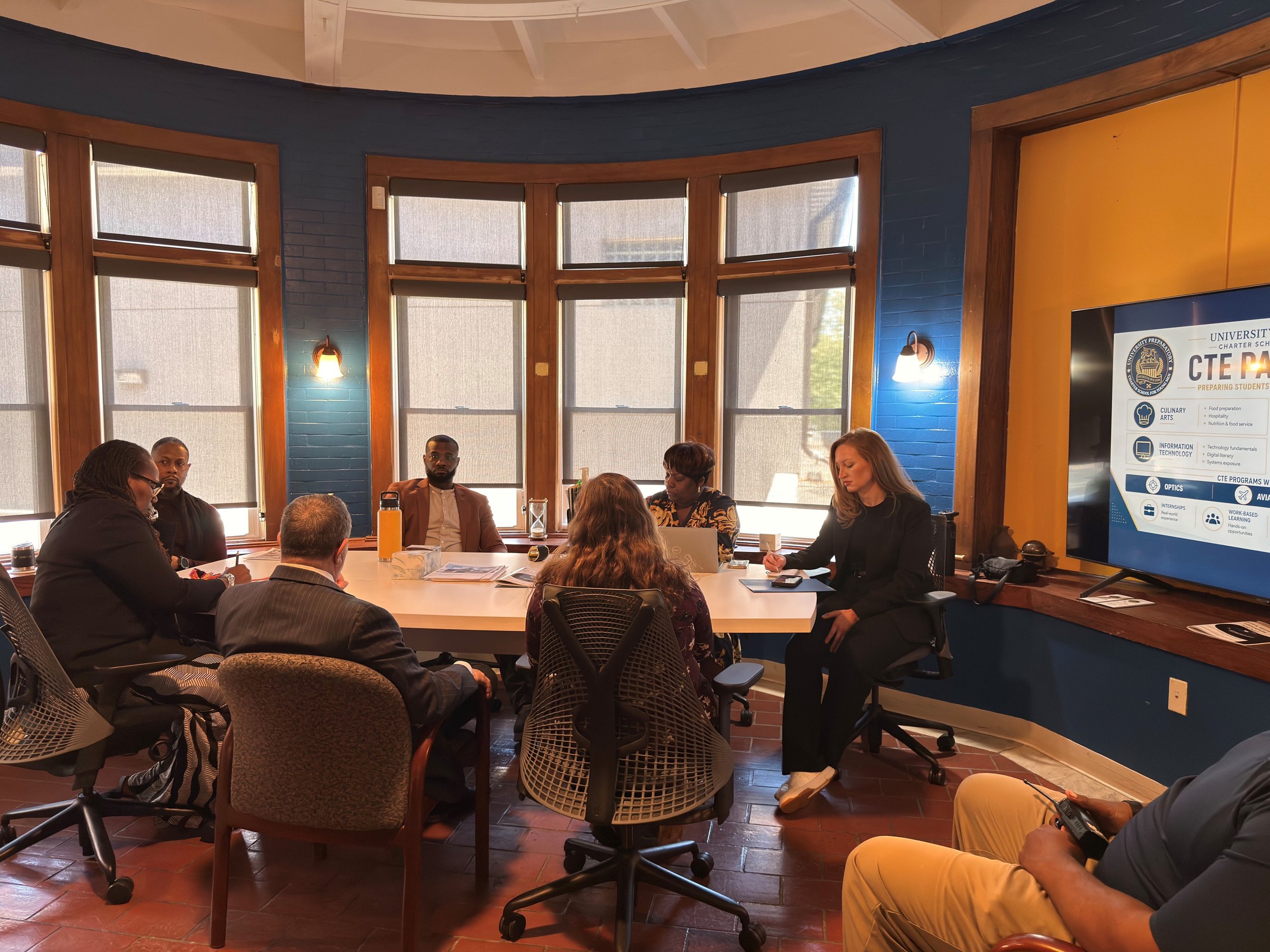 People seated around a conference table in a room with large windows, some taking notes, with a screen displaying a presentation on the right side.