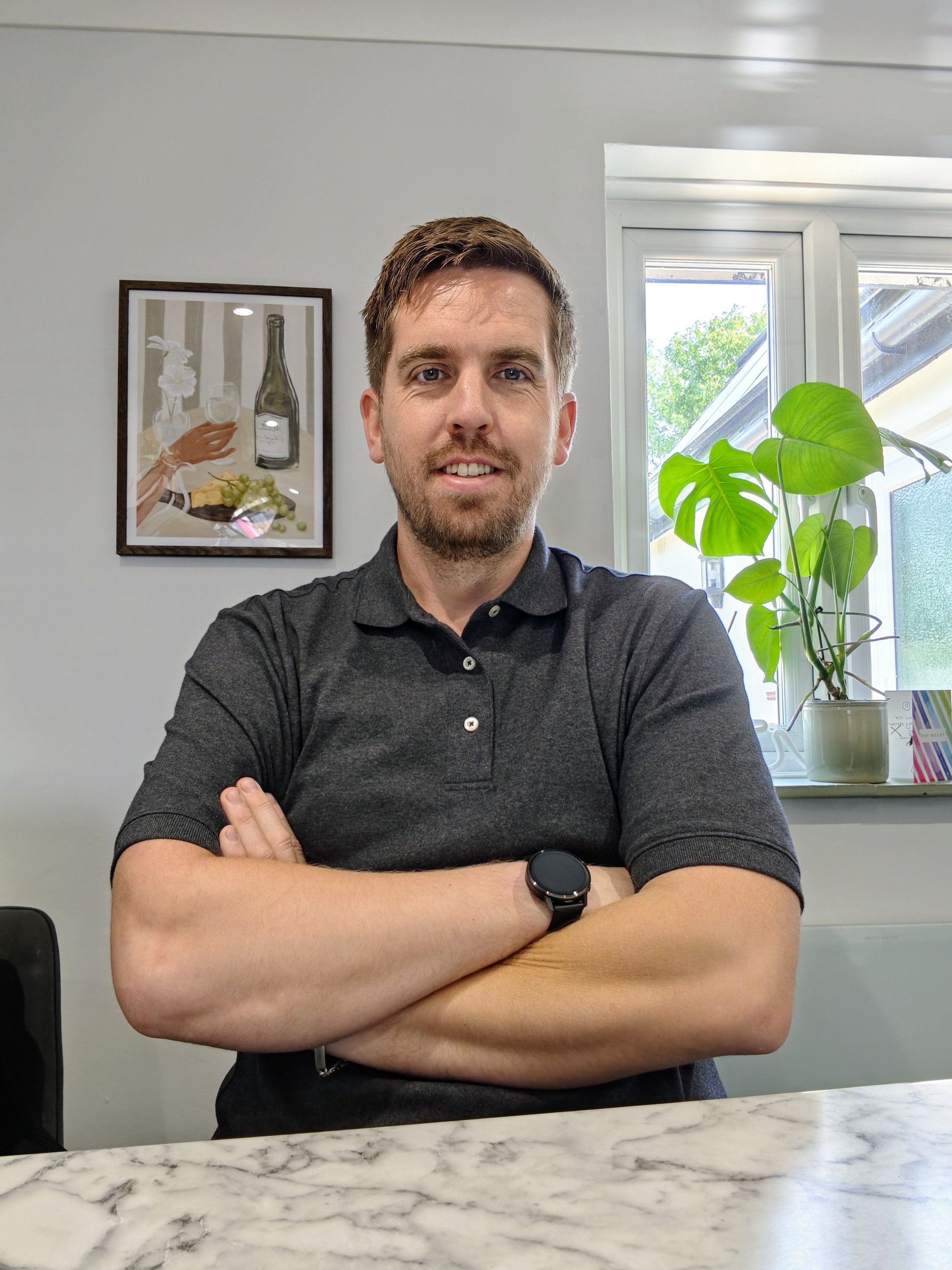 A man with brown hair and a beard, wearing a dark polo shirt and a smartwatch, sitting with arms crossed at a marble table in a room with a window, a green plant, and framed artwork in the background.