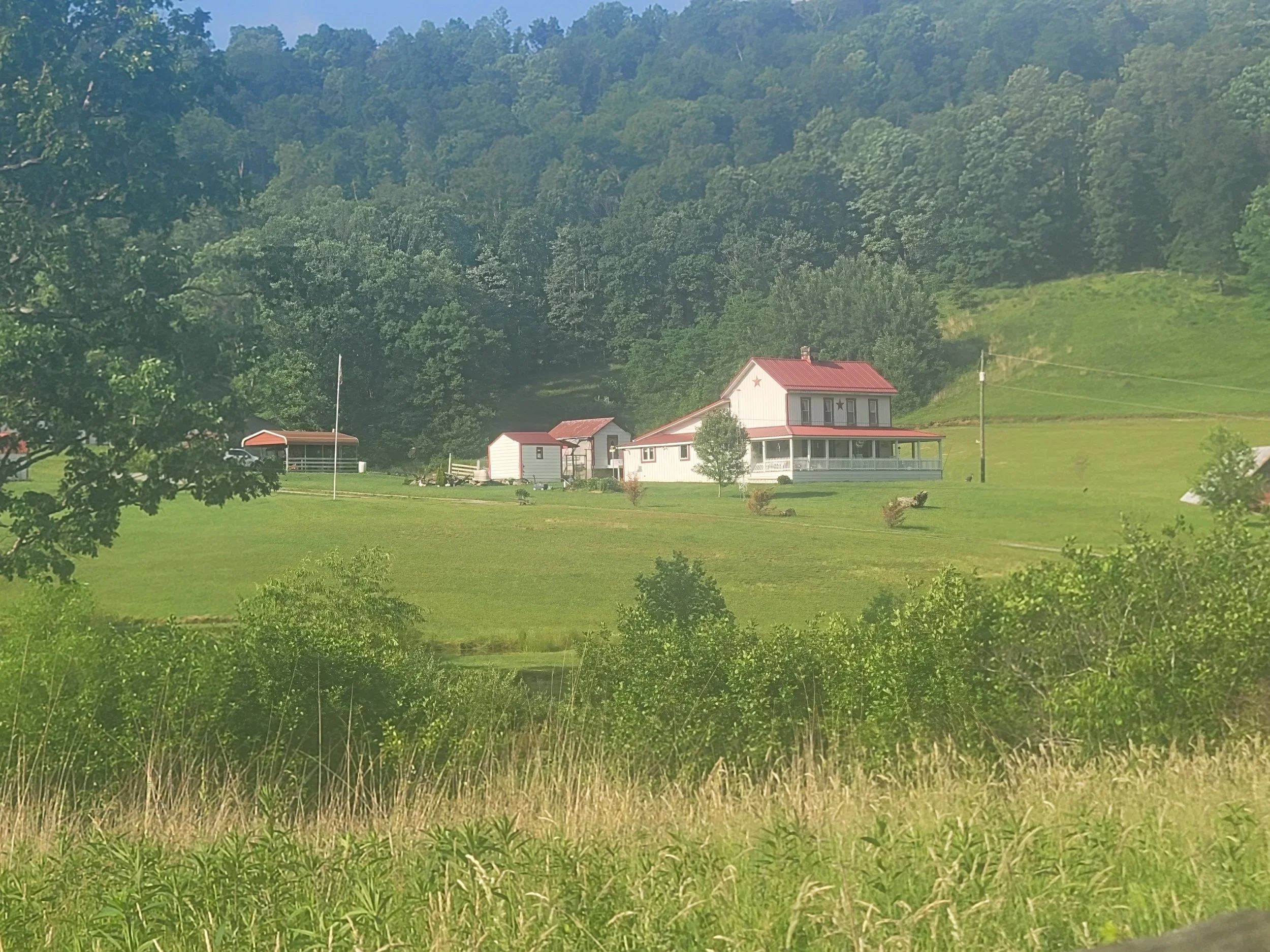A scenic rural landscape featuring green rolling hills, a white house with a red roof, and dense trees on the hillside in the background.