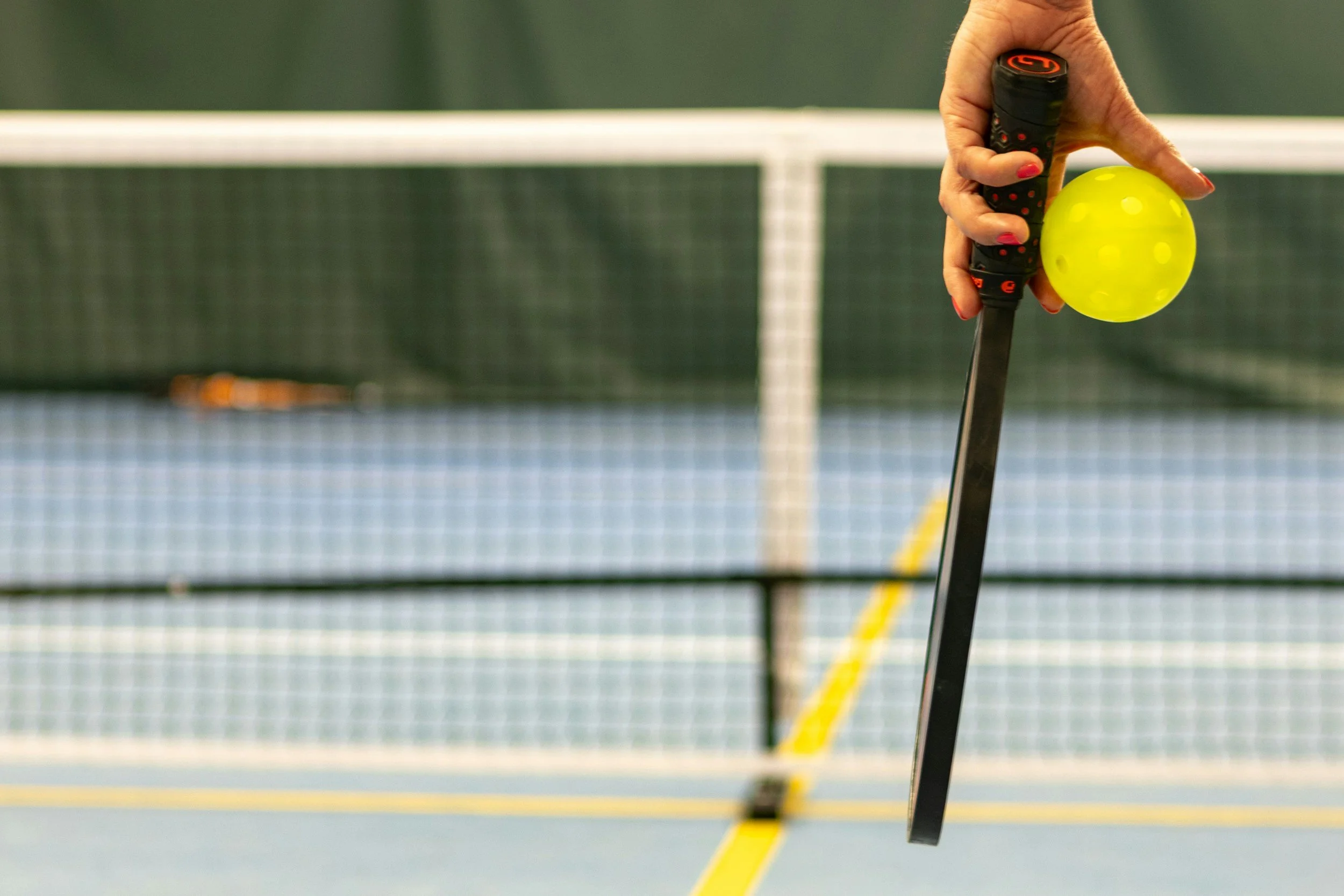 Close-up of a hand holding a yellow  pickleball and a black paddle during a pickleball game on an indoor court.