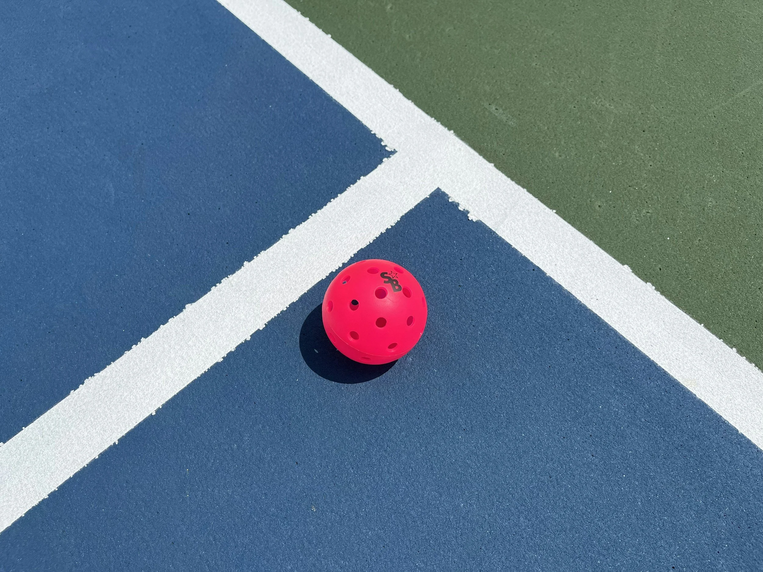 A close-up of a pink pickleball on a blue and green pickleball court with white boundary lines.