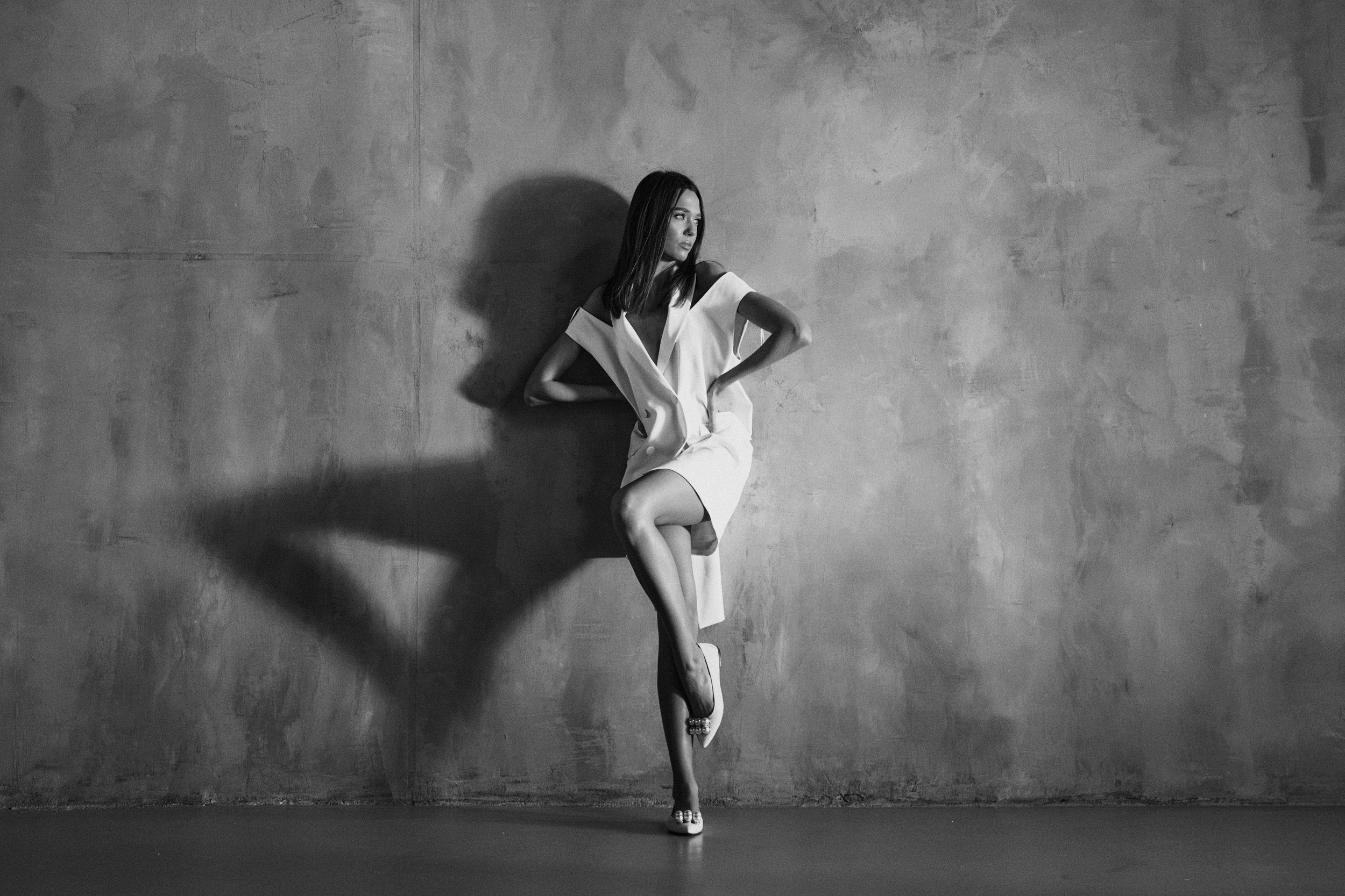 Black-and-white fashion portrait of a woman in a structured white dress and heels, posed against a textured wall with a dramatic elongated shadow.
