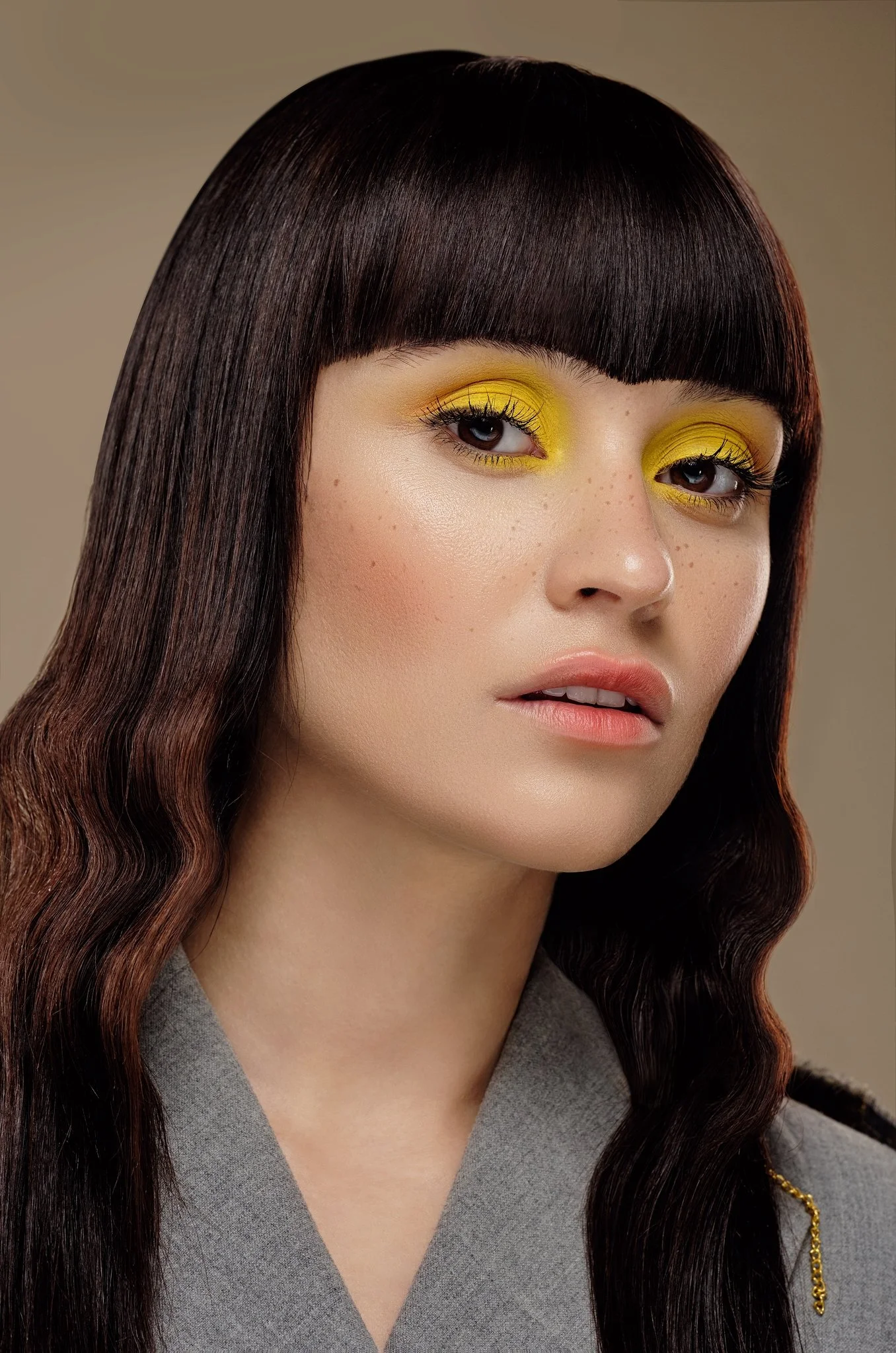 Editorial beauty portrait featuring vivid yellow eye makeup, freckles, glossy skin and dark hair styled with blunt bangs and soft waves.
