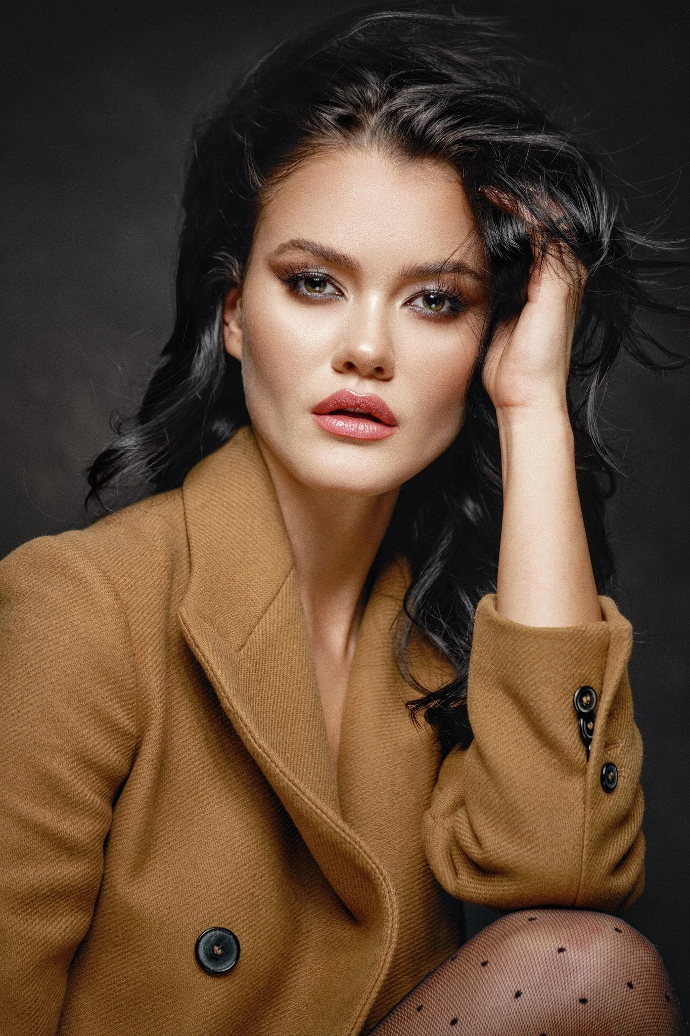 Close-up beauty portrait of a dark-haired woman with luminous makeup, glossy pink lips and a camel tailored blazer.
