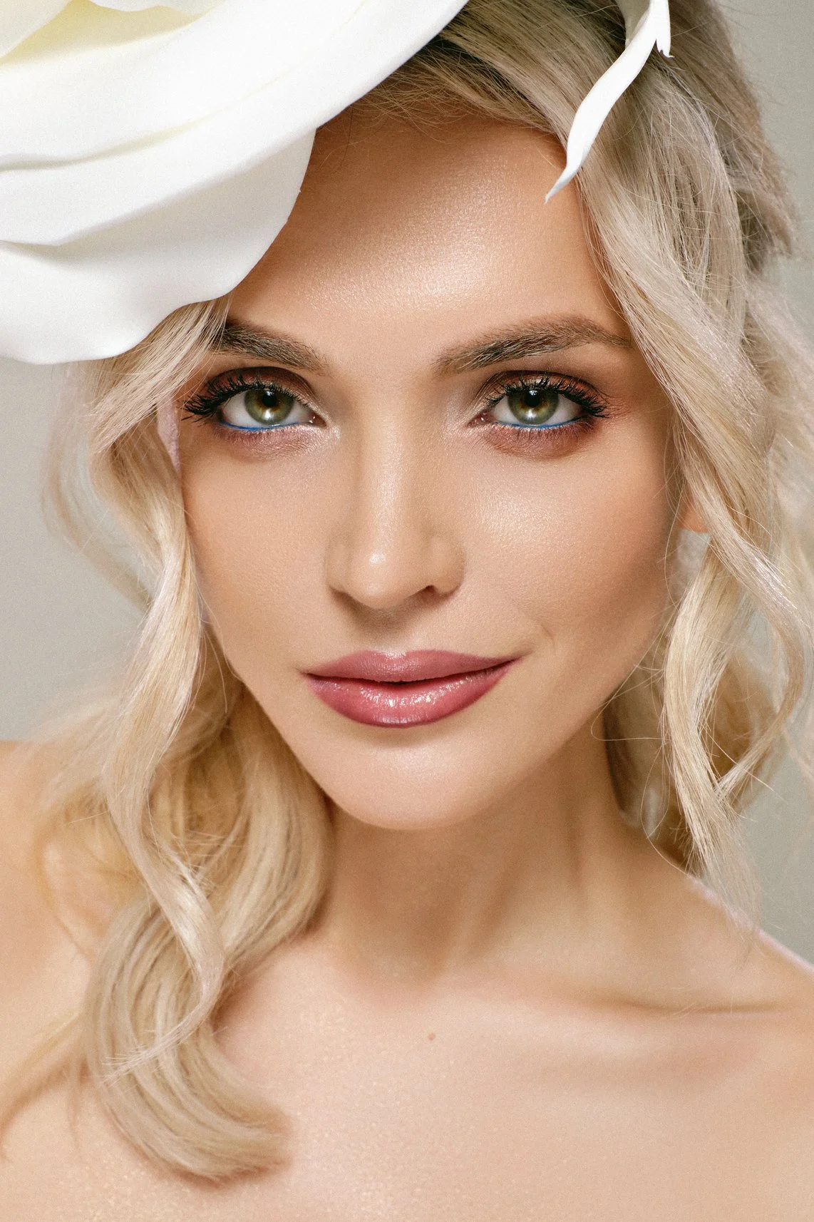 Luminous beauty portrait of a blonde woman with green eyes, soft pink makeup and an oversized white flower framing her face.
