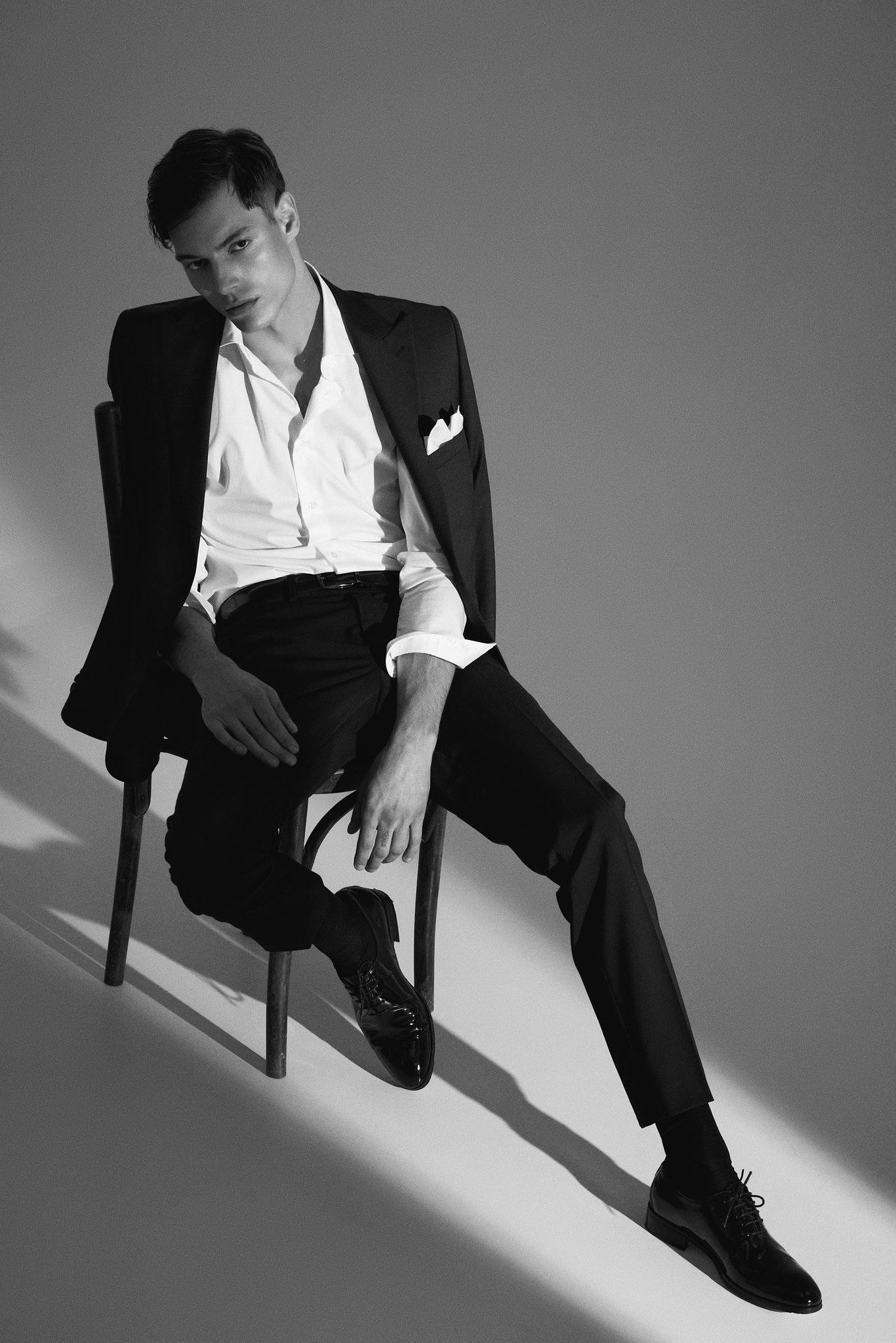Full-length black-and-white fashion portrait of a model seated in a black suit and white shirt under dramatic directional light.
