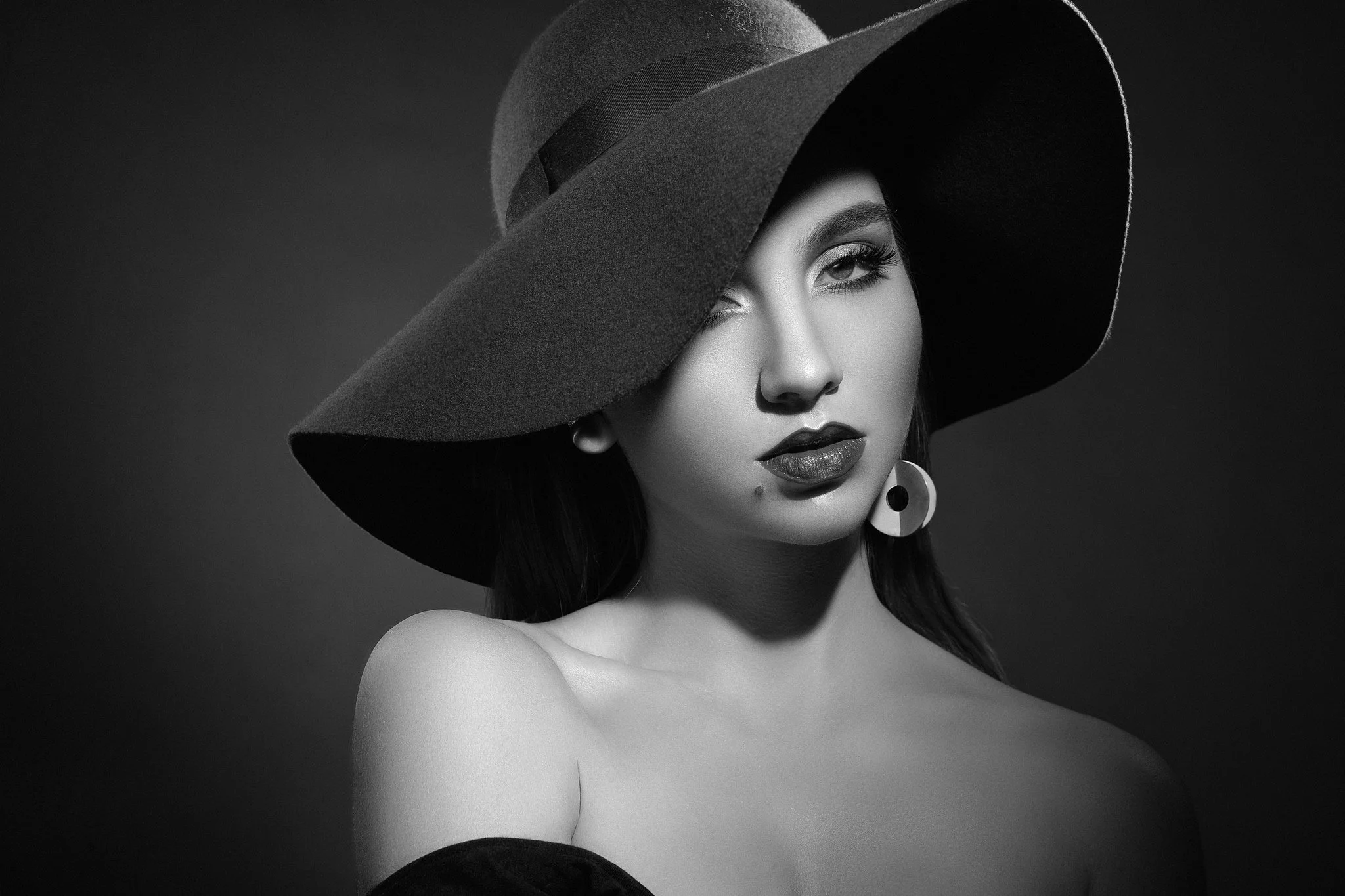 Black-and-white fashion portrait of a woman wearing a wide-brim hat, bold lipstick and a circular statement earring.
