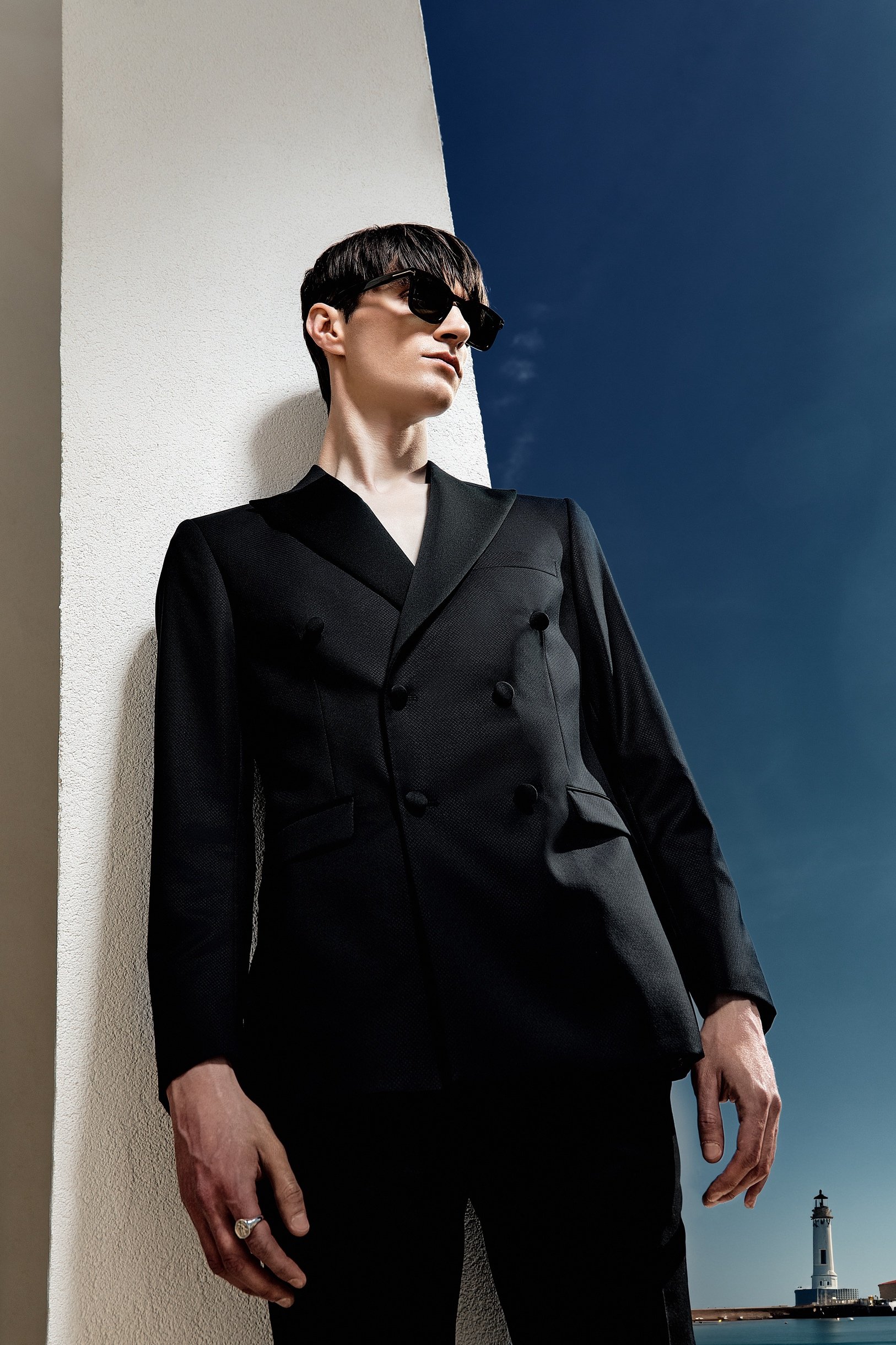Low-angle menswear portrait of a model in a black double-breasted suit and sunglasses against a deep blue sky and coastal lighthouse.
