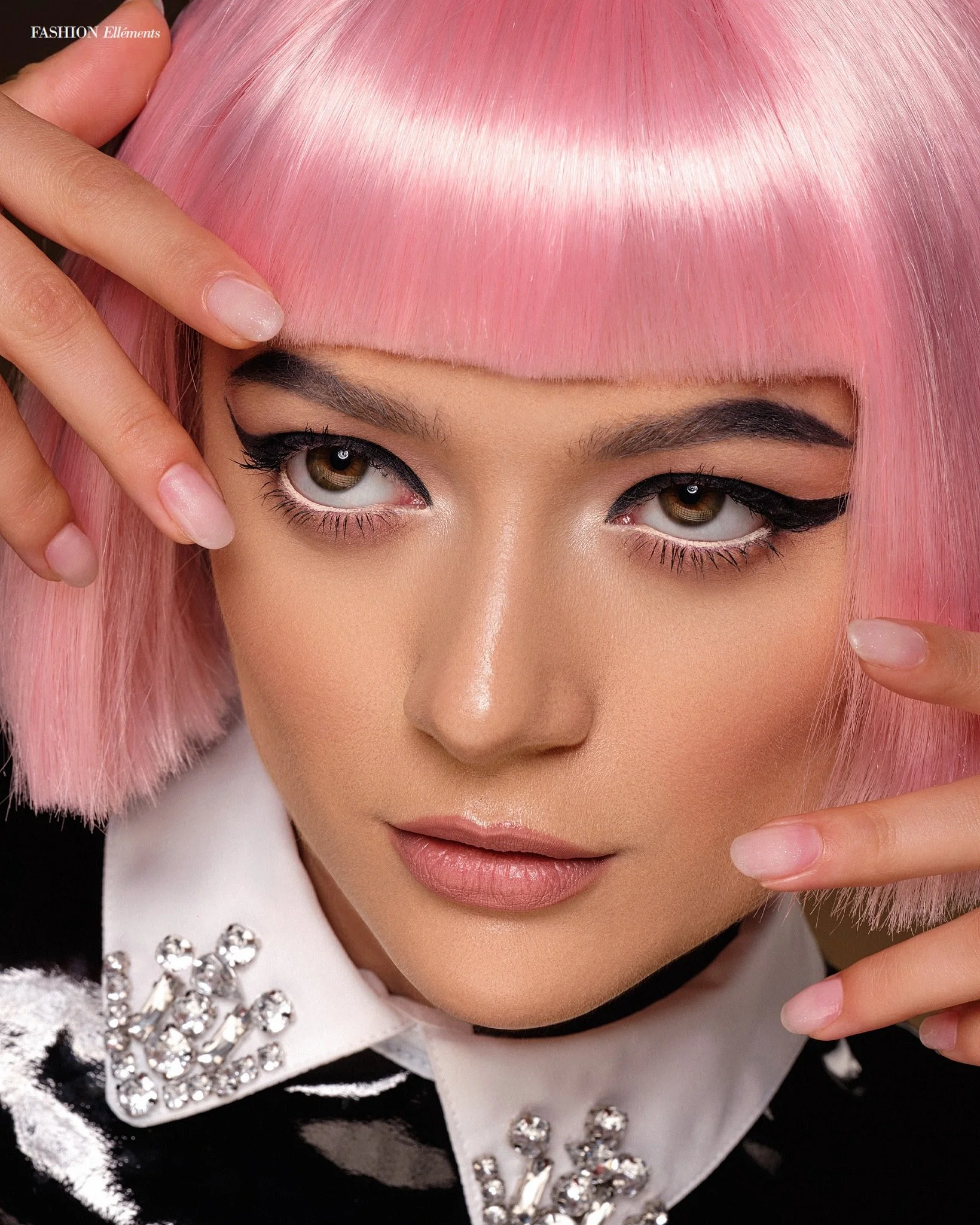 Close-up beauty editorial featuring a model with a glossy pink bob, graphic black eyeliner, manicured hands and an embellished white collar.
