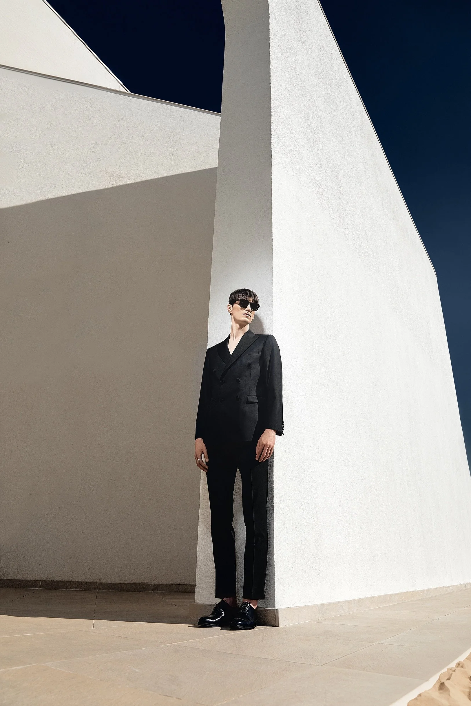 Full-length menswear campaign portrait of a model in a black double-breasted suit and sunglasses against modern white architecture.
