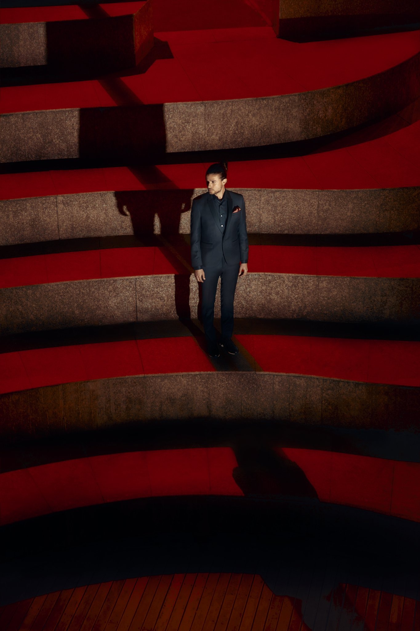 Menswear campaign portrait of a man in a dark tailored suit standing on curved red architectural steps under dramatic sunlight and elongated shadows.
