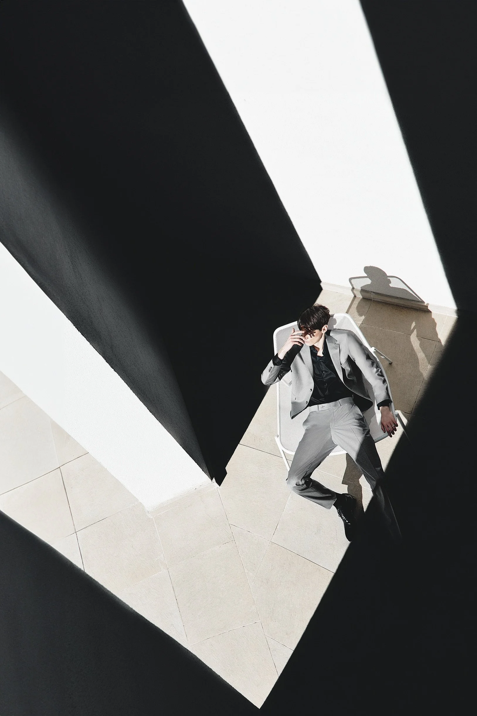 Overhead menswear campaign image of a model in a grey suit seated within bold black-and-white architectural shadows.
