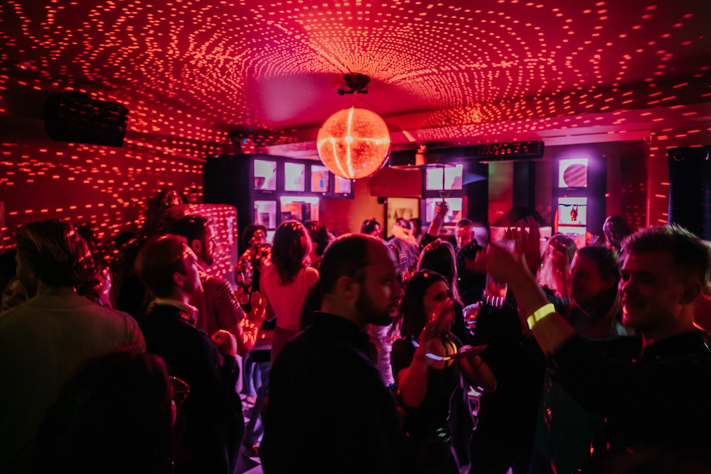 Crowd of people dancing at a What Does Not house party with red lighting, a disco ball on the ceiling, and colorful screens on the wall.